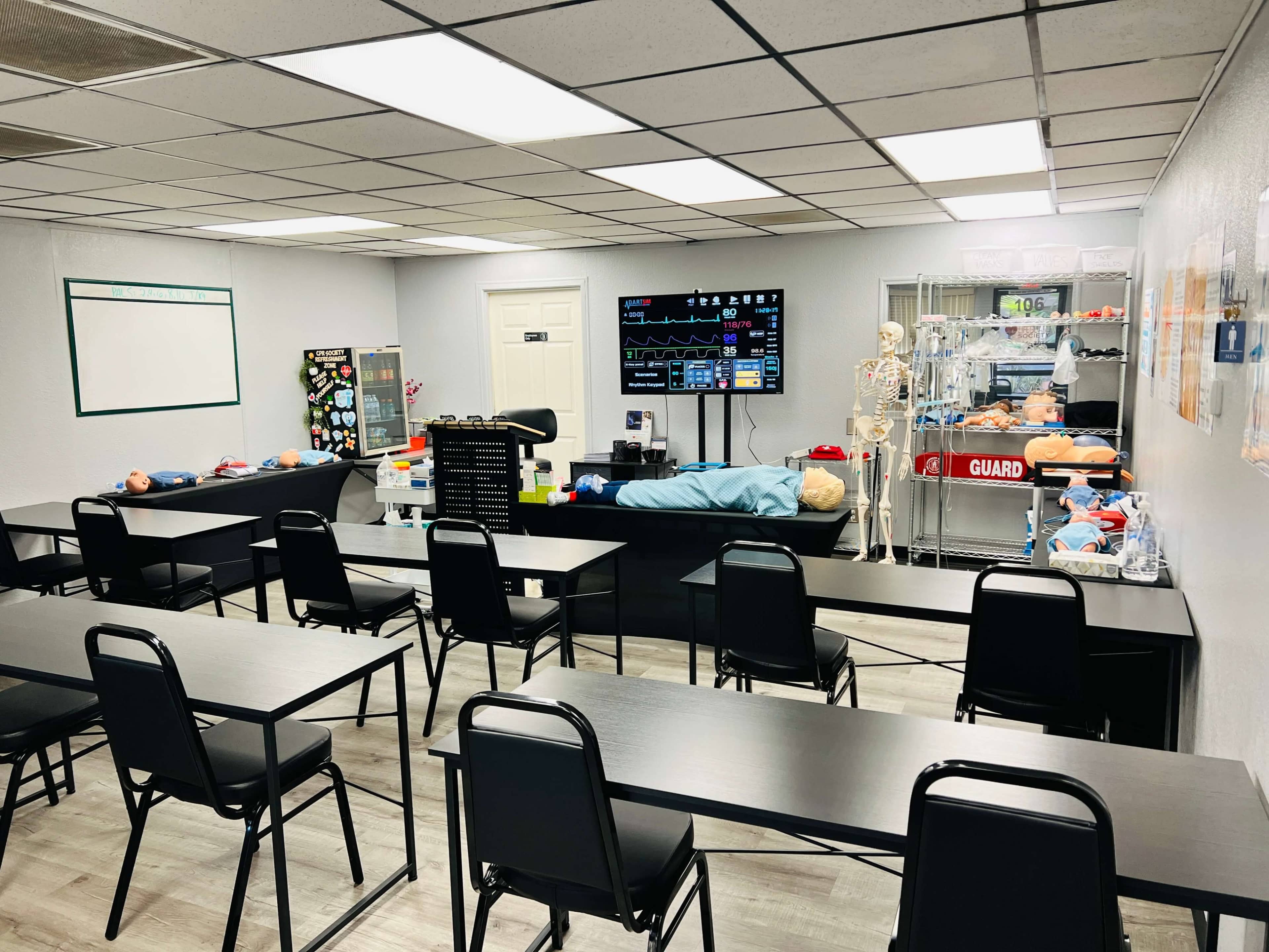 The image shows a training classroom equipped with medical mannequins, a monitor displaying vital signs, and rows of black tables and chairs.