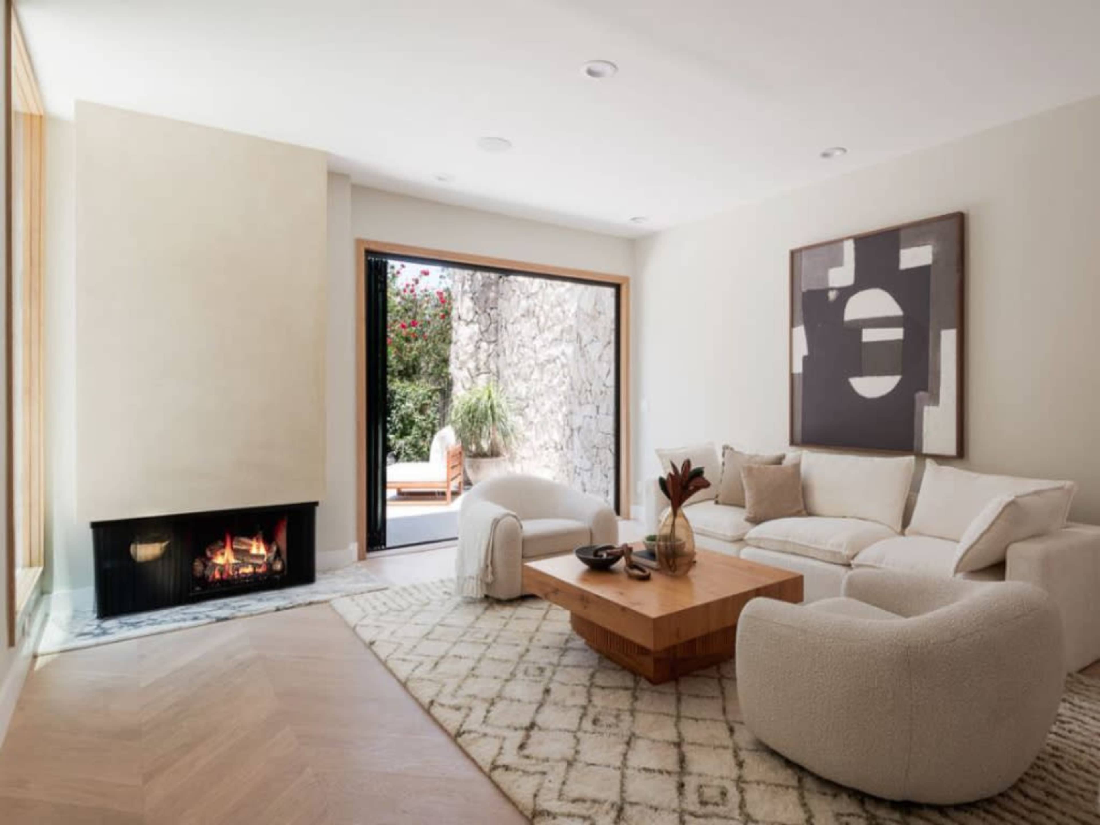 A modern living room features a curved sofa, a wooden coffee table, and a fireplace, with a view of an outdoor space through an open door.