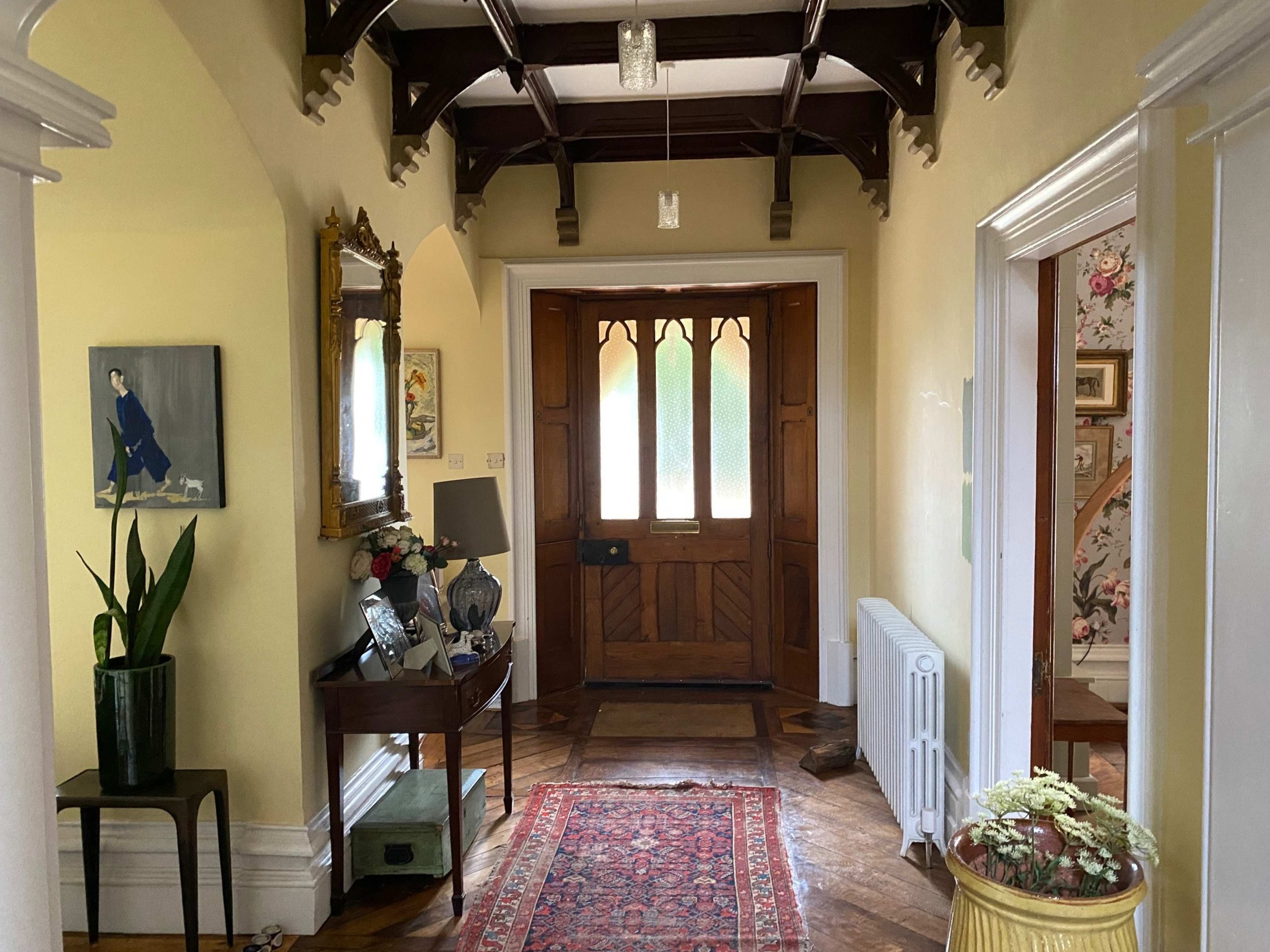 A hallway with a wooden front door, a decorative mirror, a plant, and a patterned rug on the wooden floor.