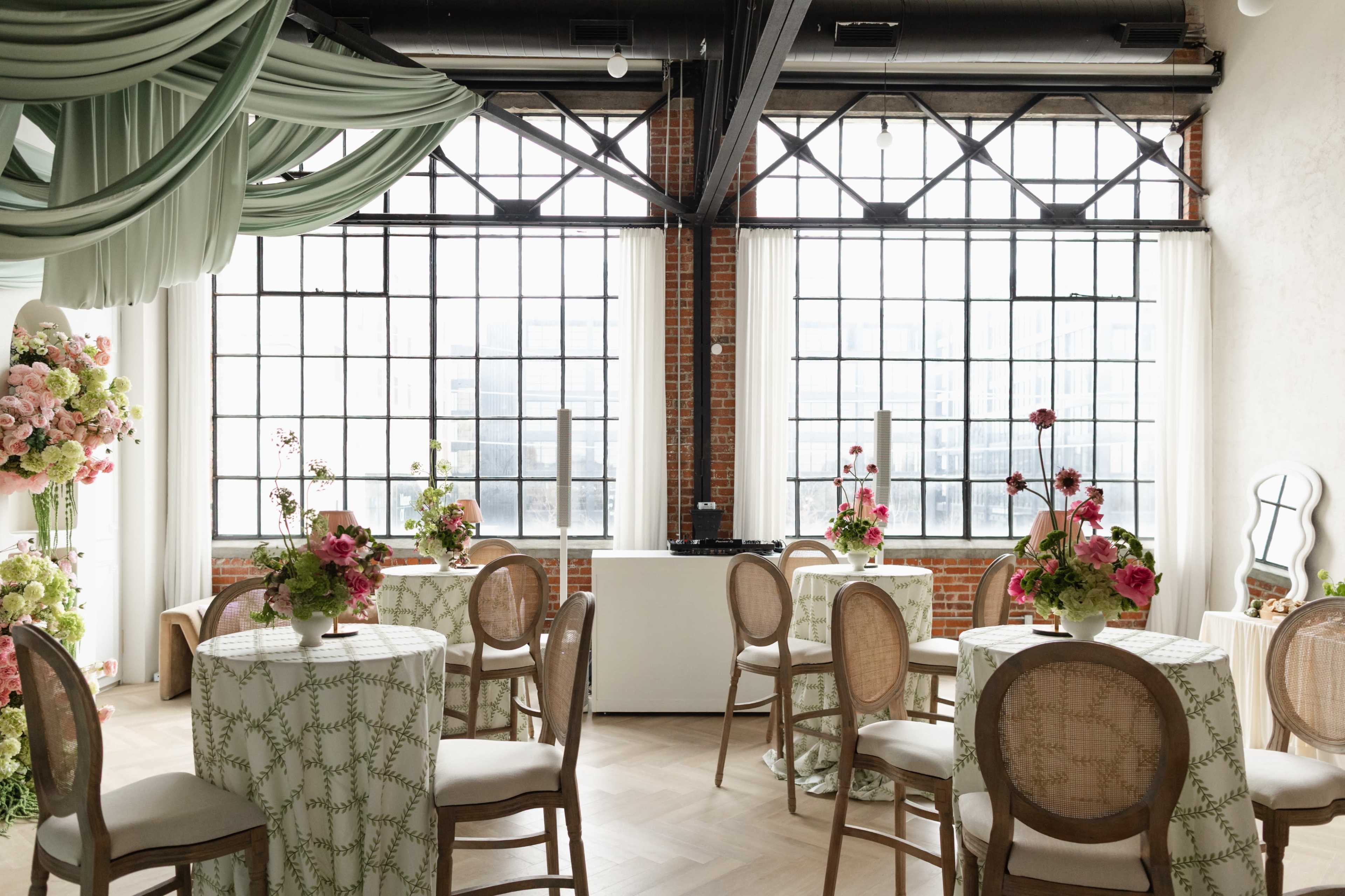 The image features a stylish event space with round tables adorned with floral centerpieces and draped fabric hanging from the ceiling, beside large industrial windows.