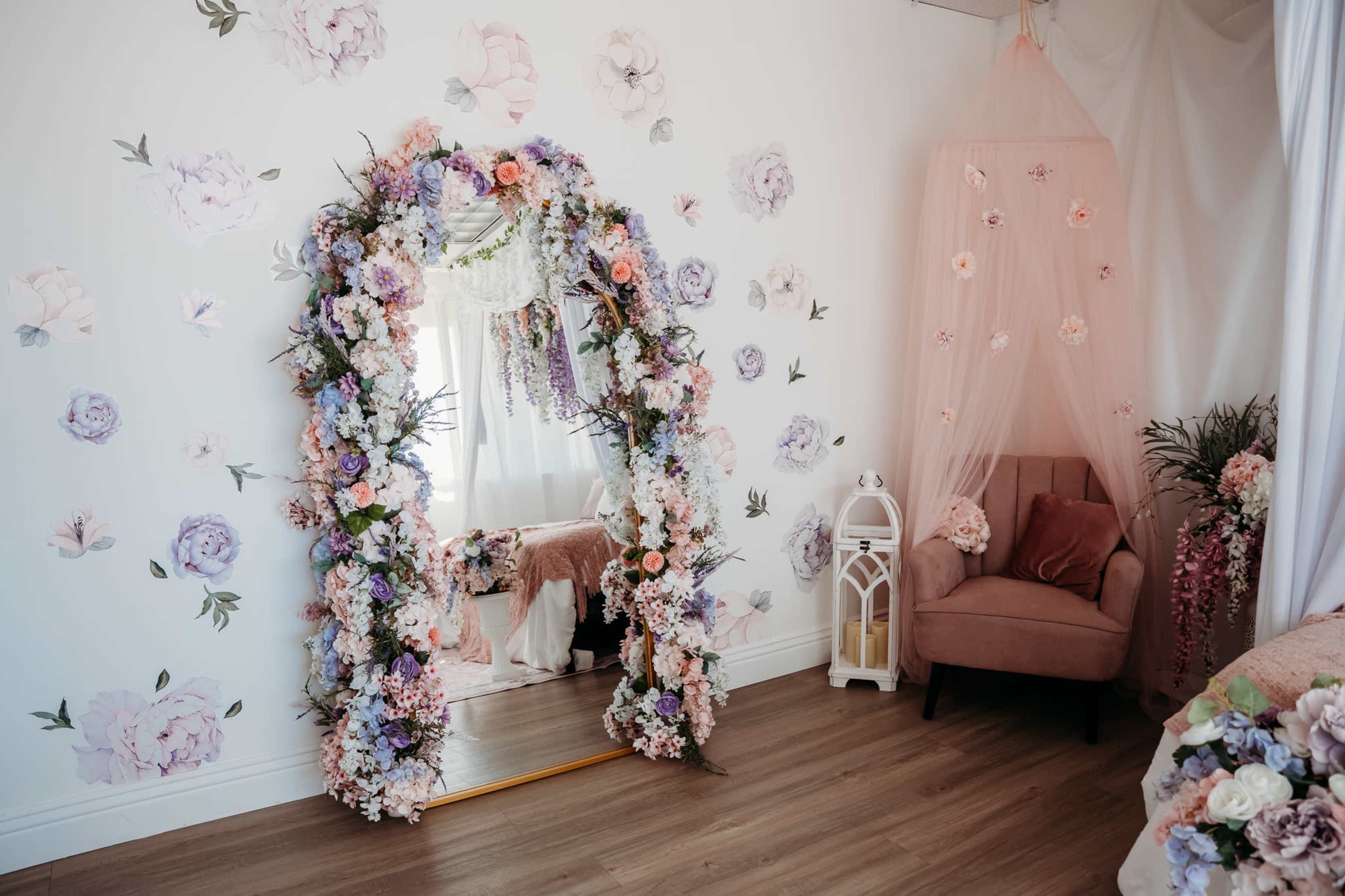 The image shows a beautifully decorated room featuring a large mirror framed with colorful artificial flowers, floral wall decals, a pink chair, and a canopy draped in tulle.