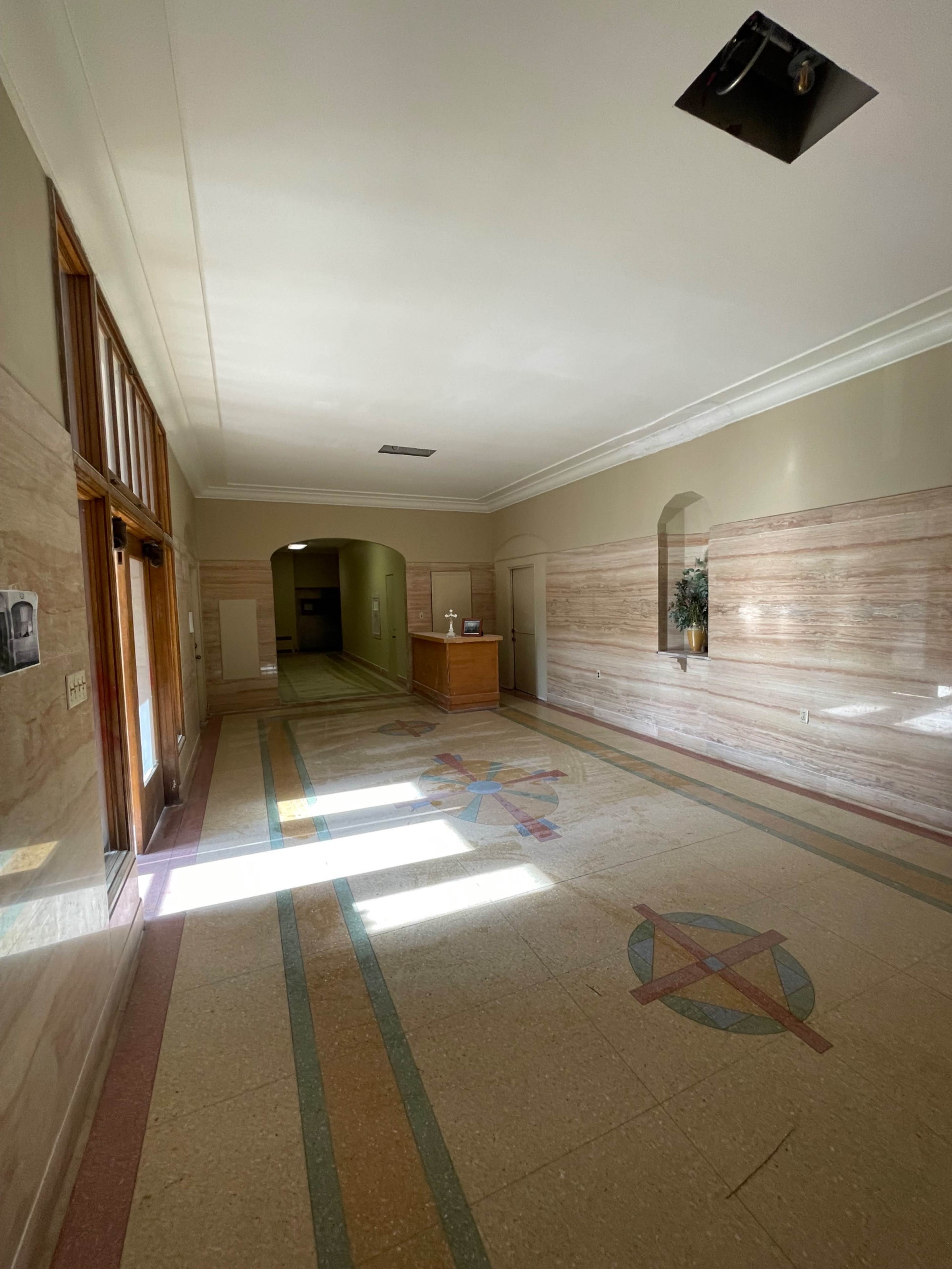 The image shows a well-lit corridor with decorative walls, a patterned floor, and a small wooden reception desk at the far end.