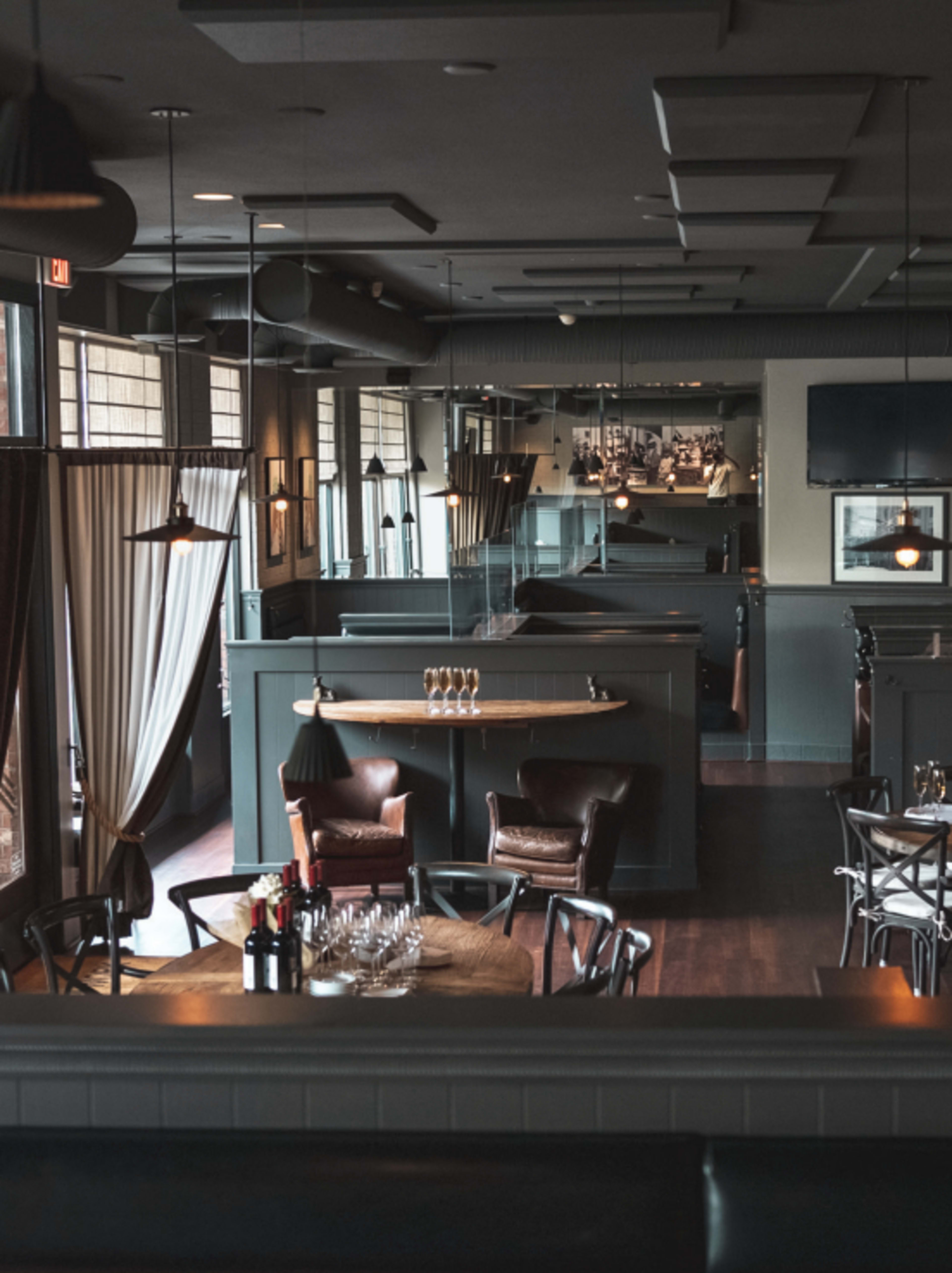 A dimly lit restaurant interior with wooden tables, leather chairs, and large windows, featuring a bar area in the background.