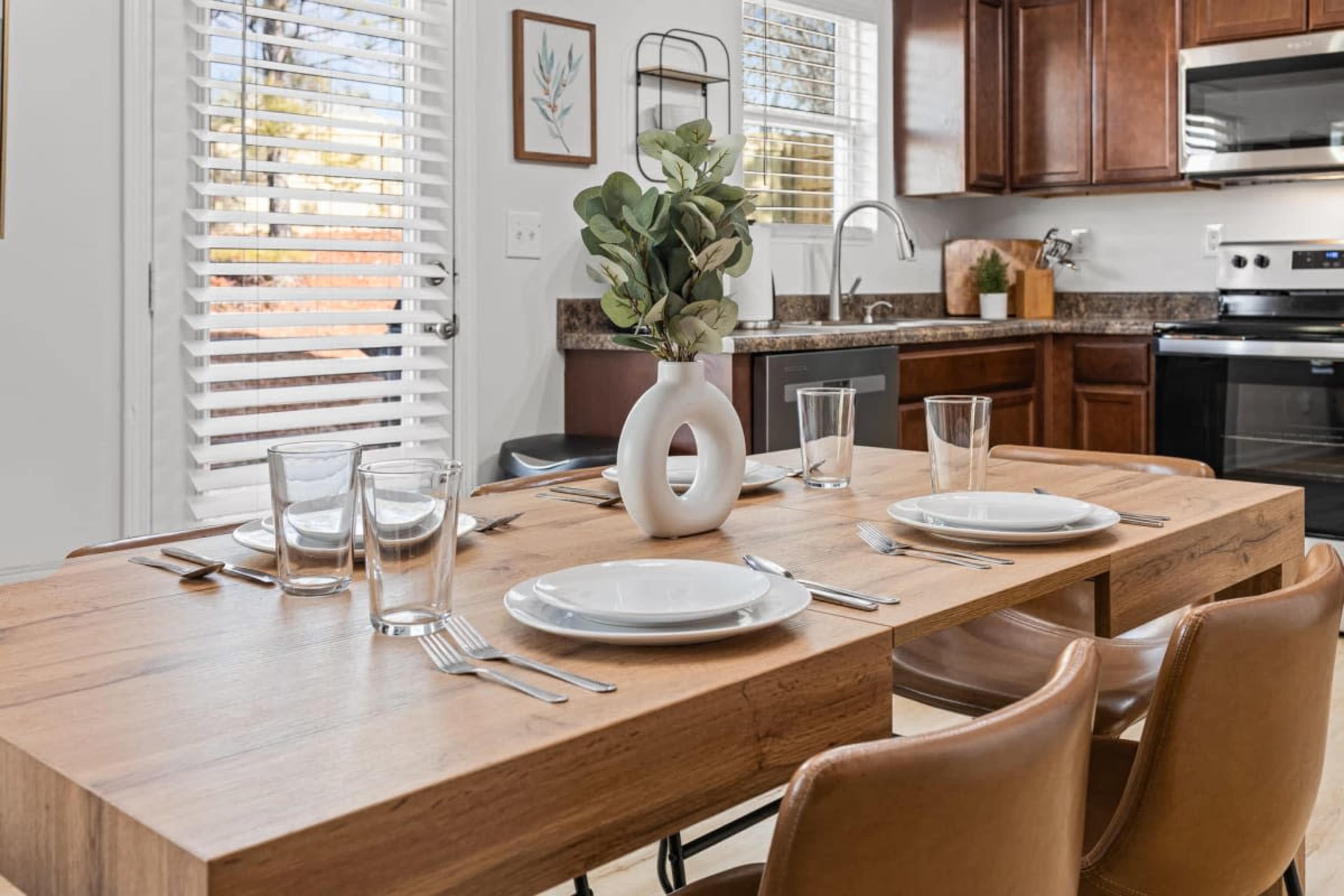 A dining table is set with plates and glasses, accompanied by a vase of greenery, in a kitchen with wooden cabinetry and ample natural light.