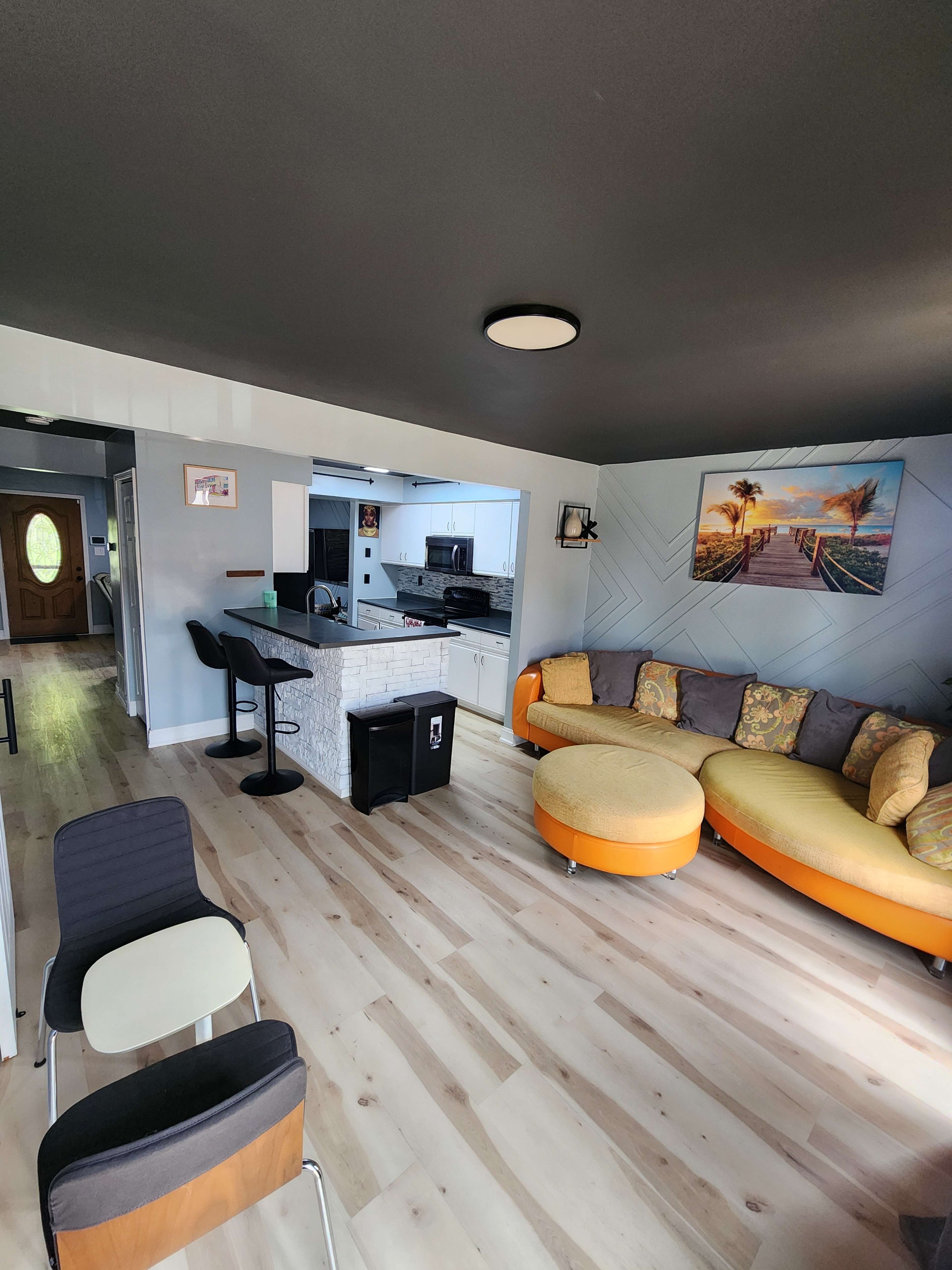 The image shows a modern living space featuring a circular orange sofa, bar stools by a kitchen island, and light-colored wooden floors.