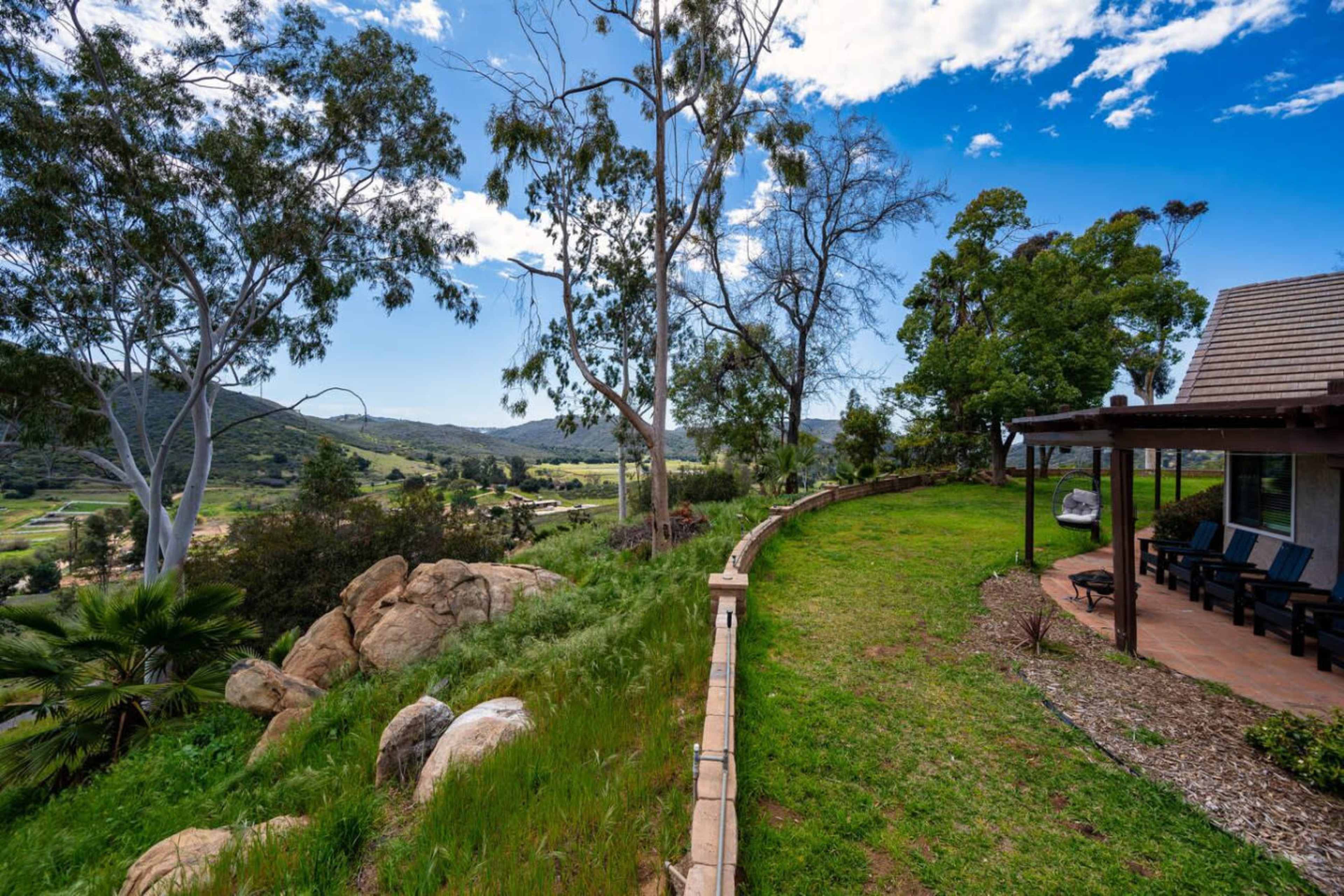 The image shows a landscaped area with a stone wall, trees, and a view of rolling hills under a partly cloudy sky.