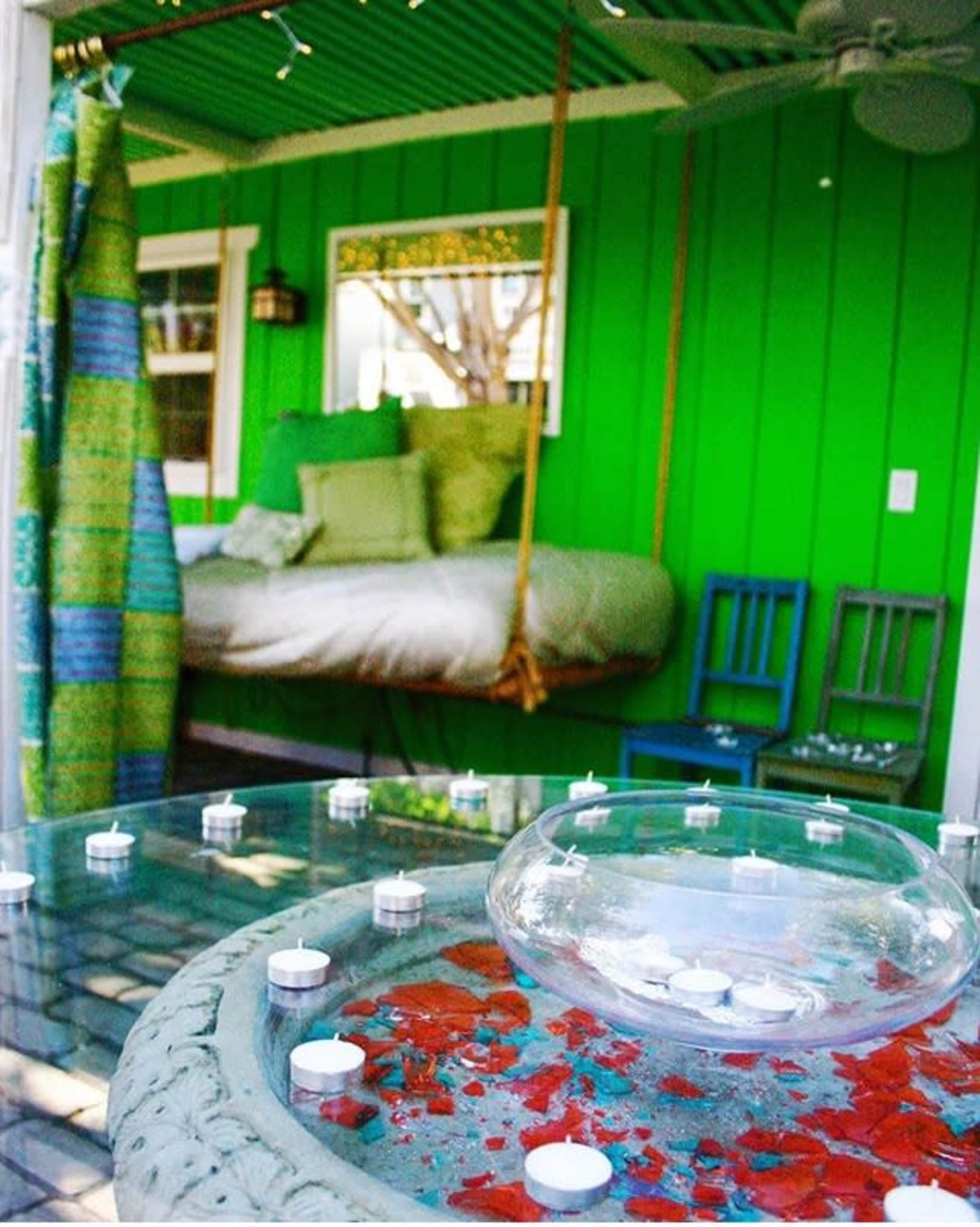 A vibrant green room with a suspended bed, a decorative table featuring a glass bowl and candles, and blue chairs.