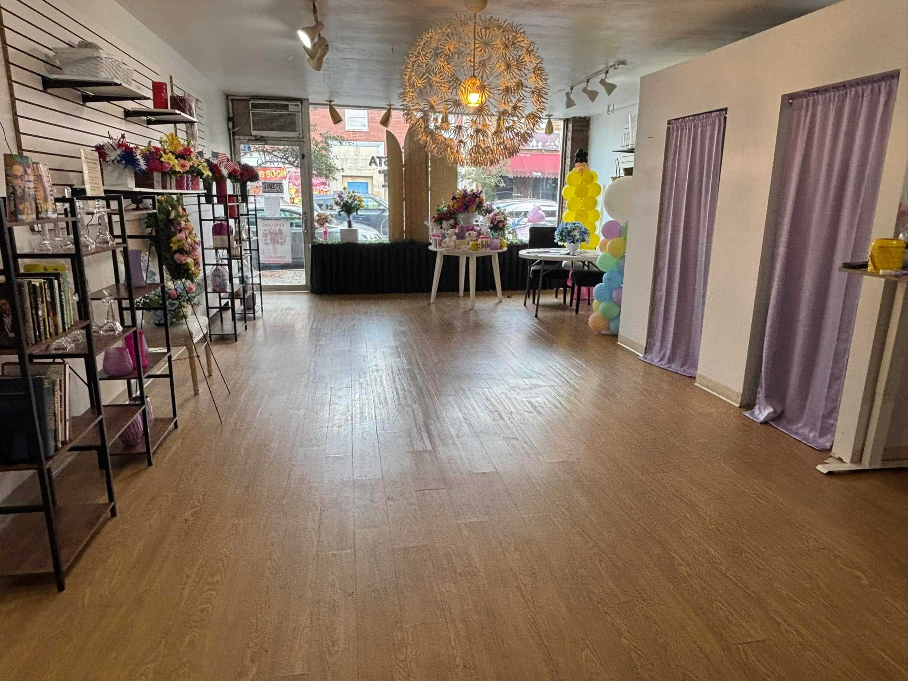 The room features wooden flooring, a large decorative light fixture, and is filled with chairs, tables, and floral arrangements against bright windows.