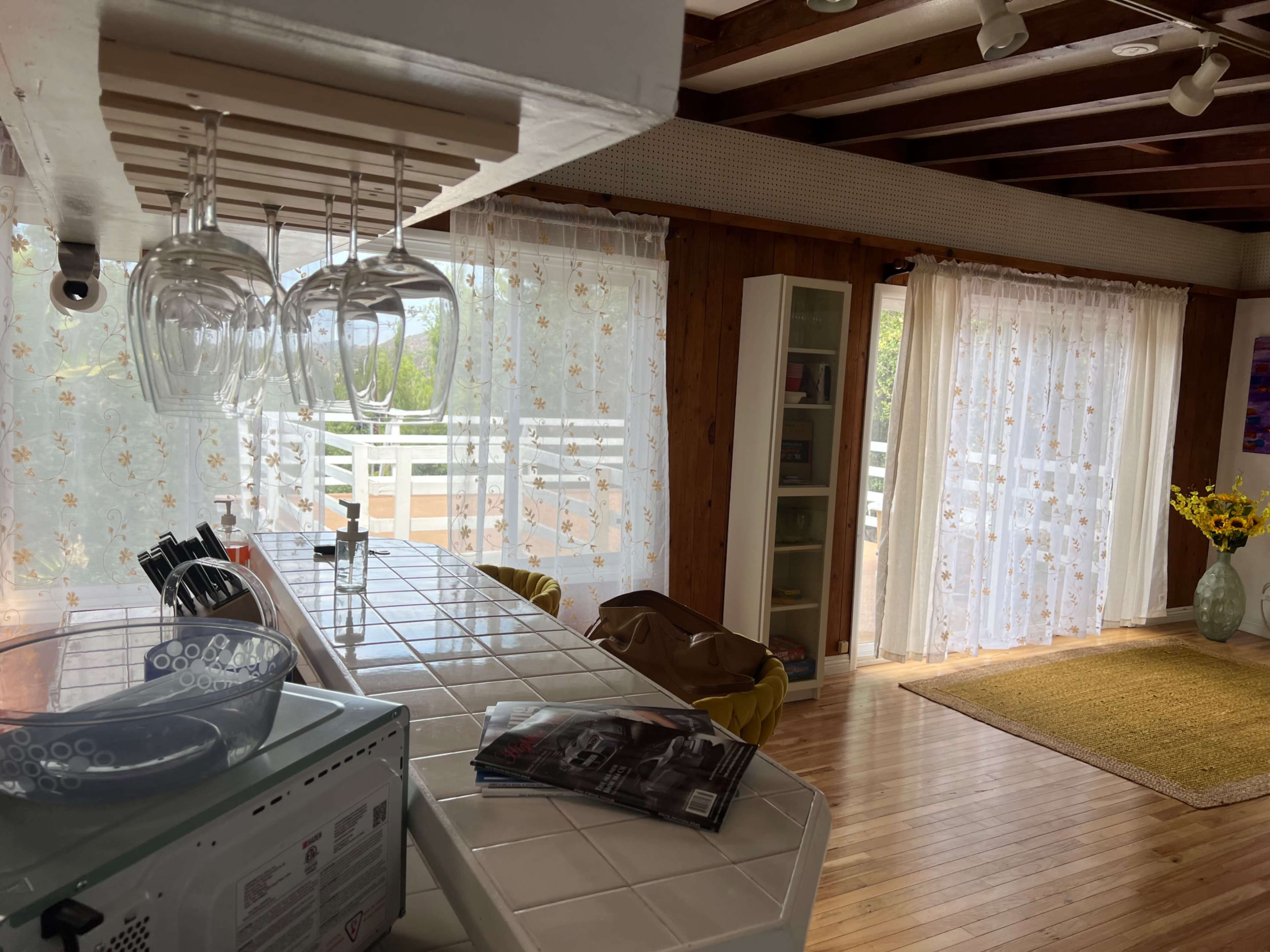 A bright kitchen area features a tiled countertop, hanging wine glasses, and large windows with sheer curtains, leading to a cozy living space.