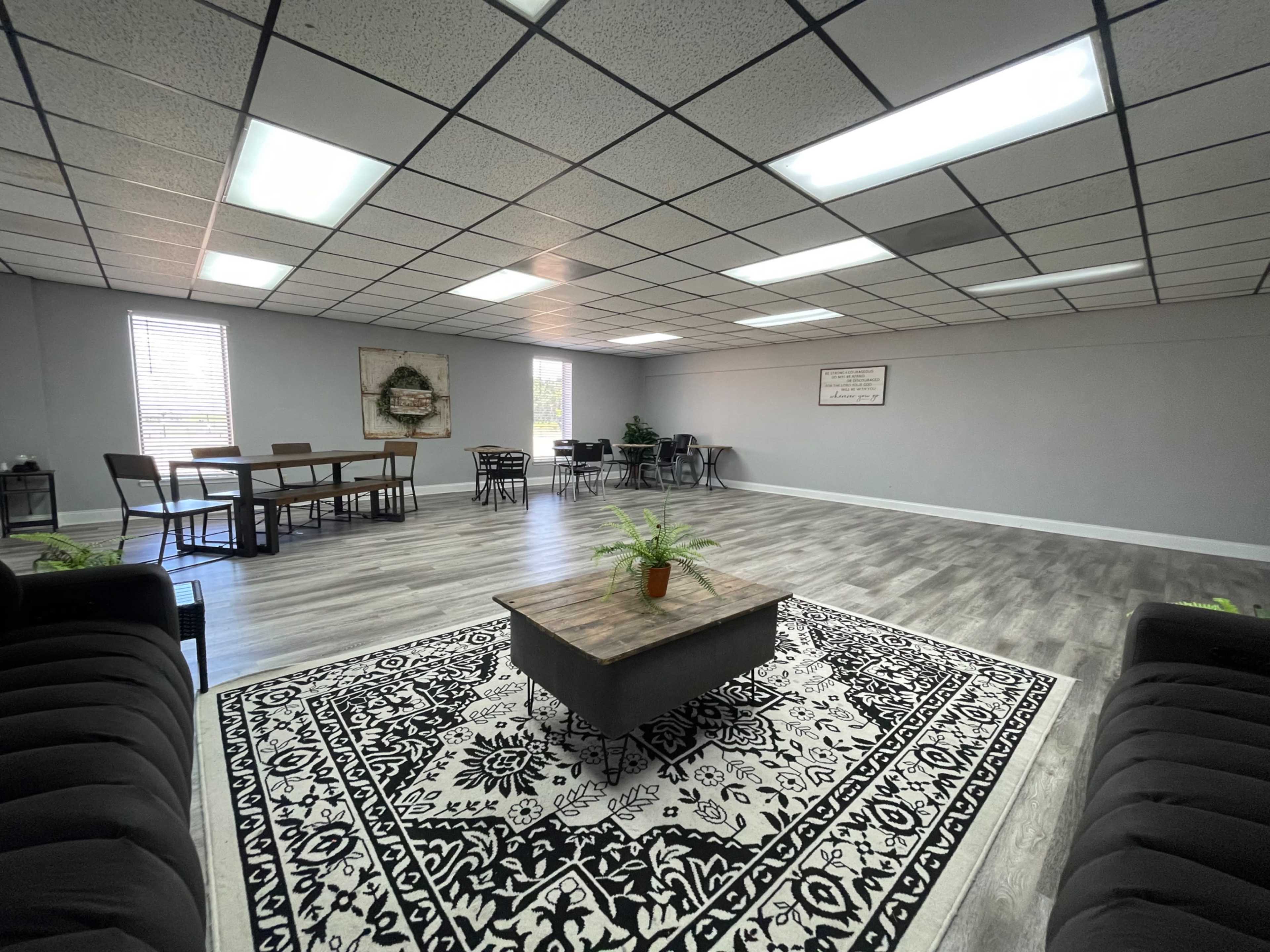 The image shows a spacious, well-lit room with a wooden table and chairs, a coffee table on a patterned rug, and several plants placed around the area.