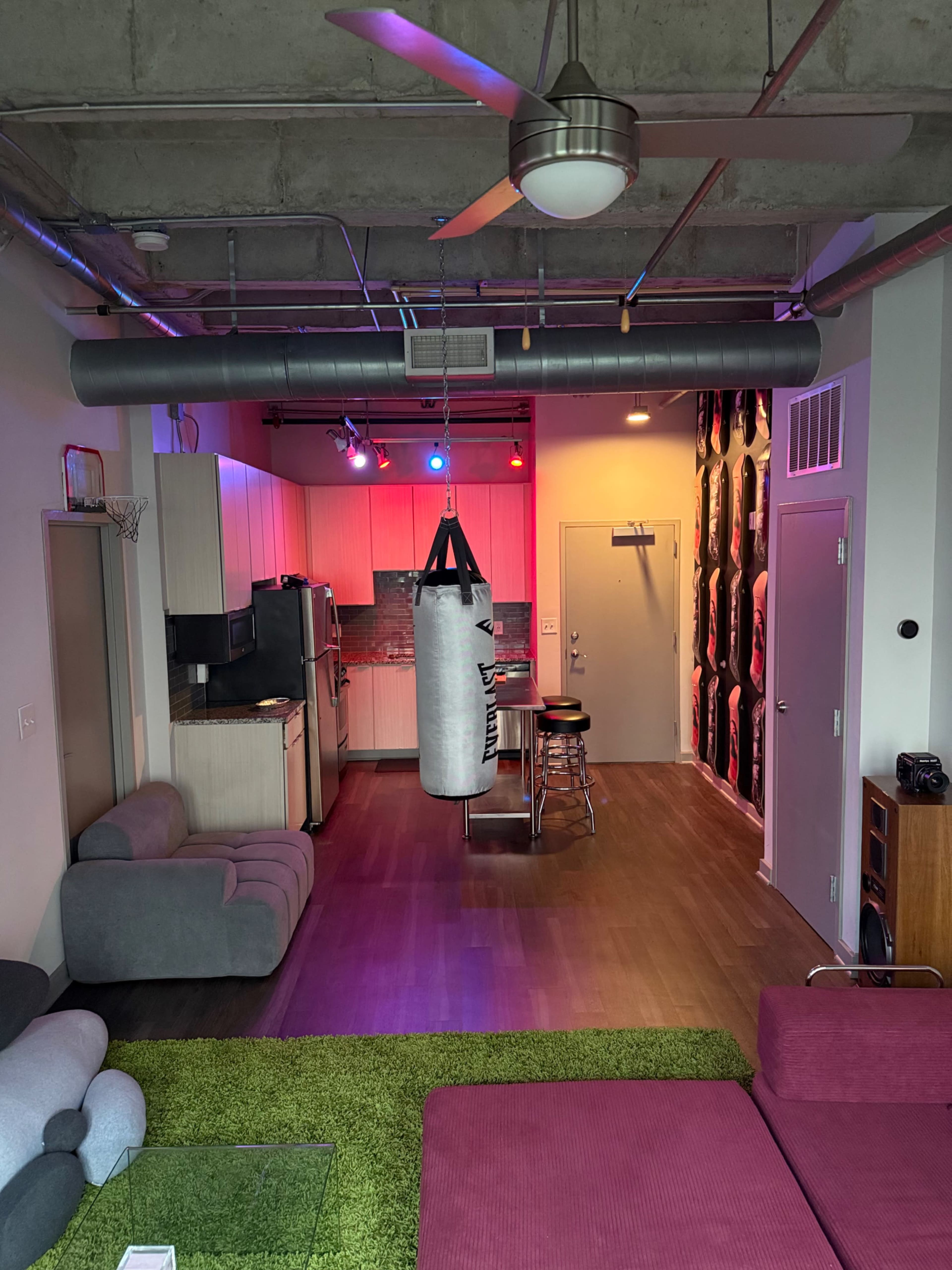 The image shows a modern, open-plan living space with a hanging punching bag, a small kitchen area, a seating area with colorful furniture, and stylish lighting.