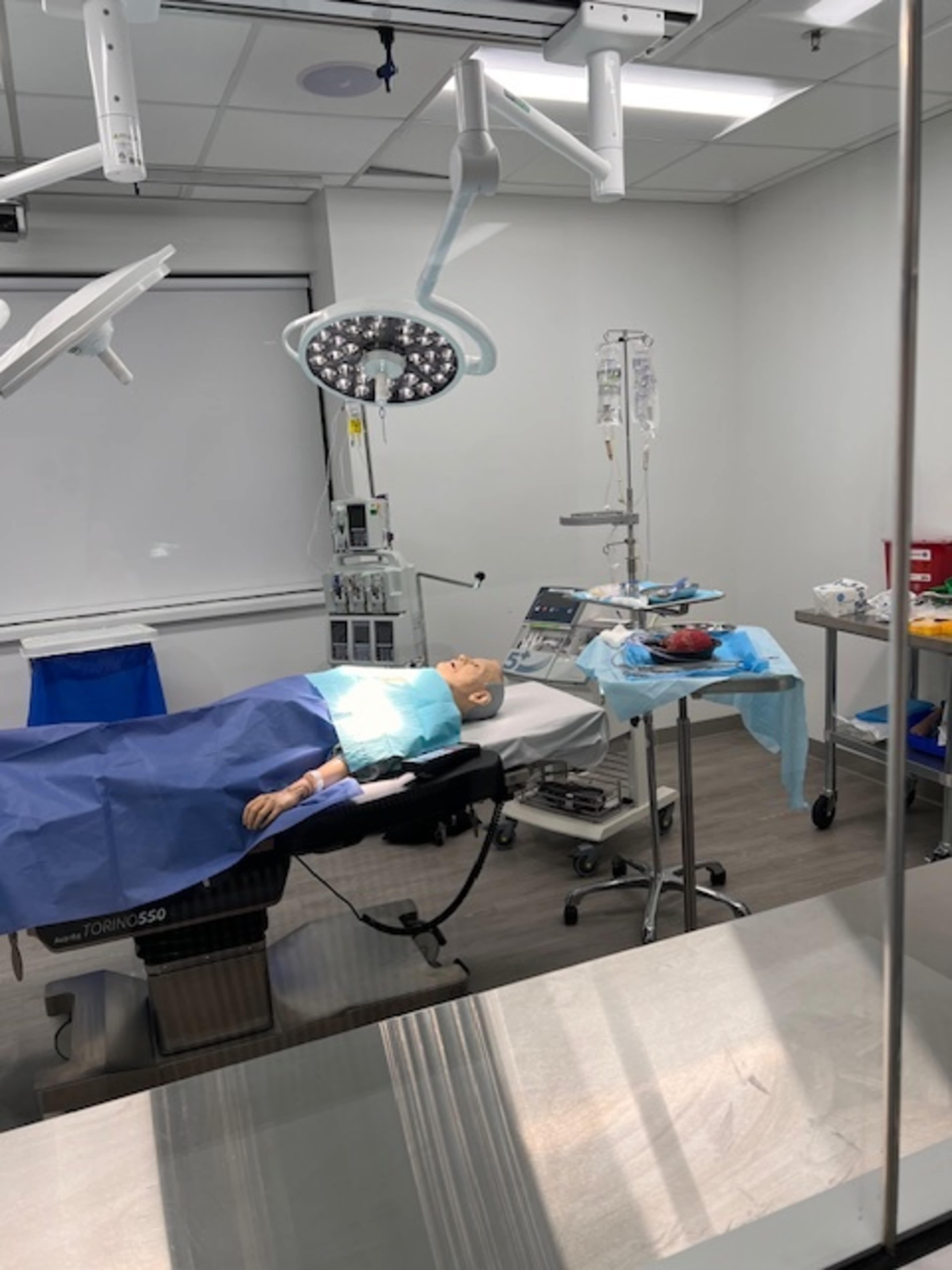 The image shows a surgical room equipped with a medical mannequin on an operating table, surrounded by various medical instruments and equipment.