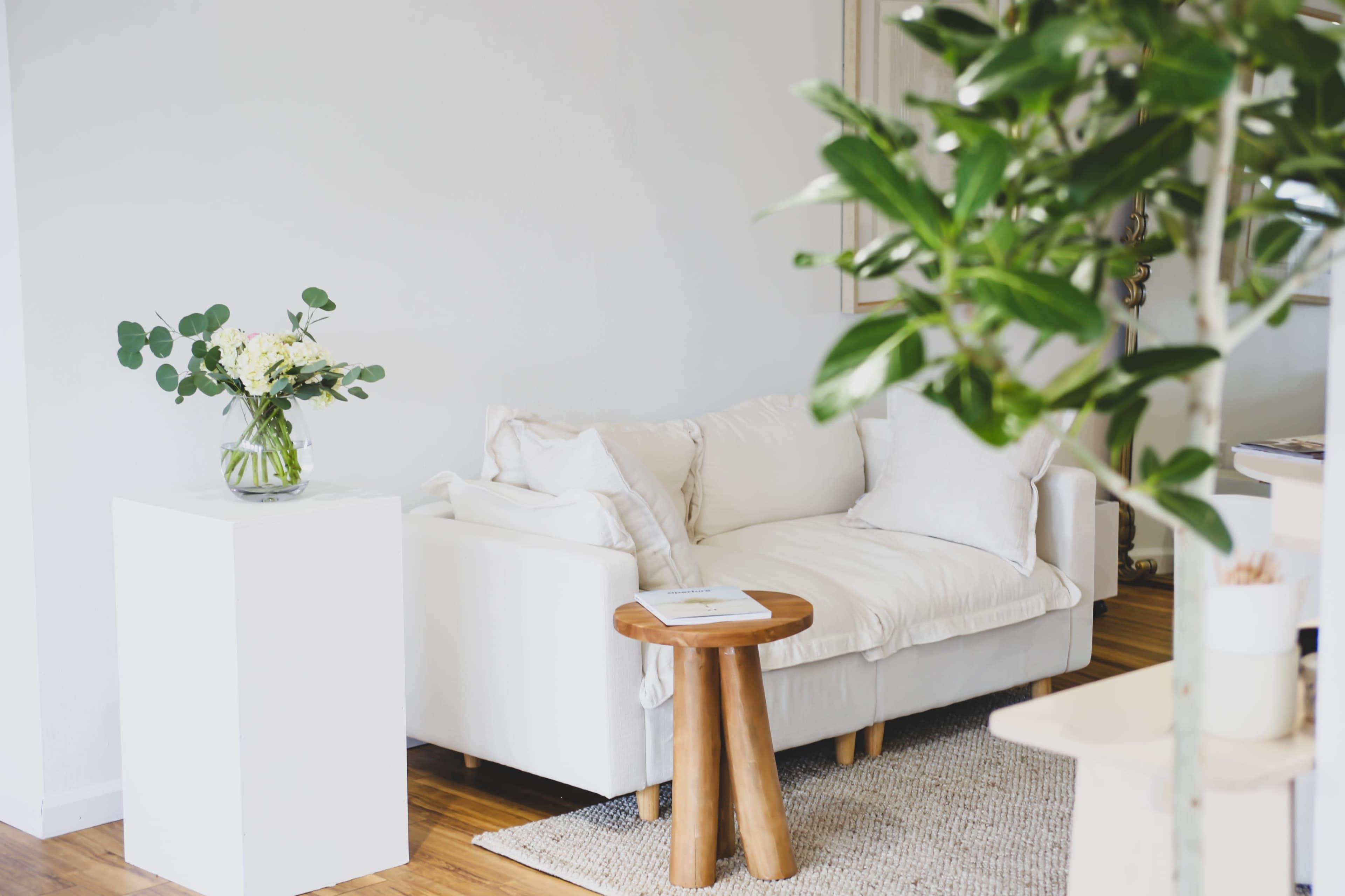 A white couch with cushions sits beside a wooden side table and a vase of flowers on a pedestal in a bright, minimalist room.