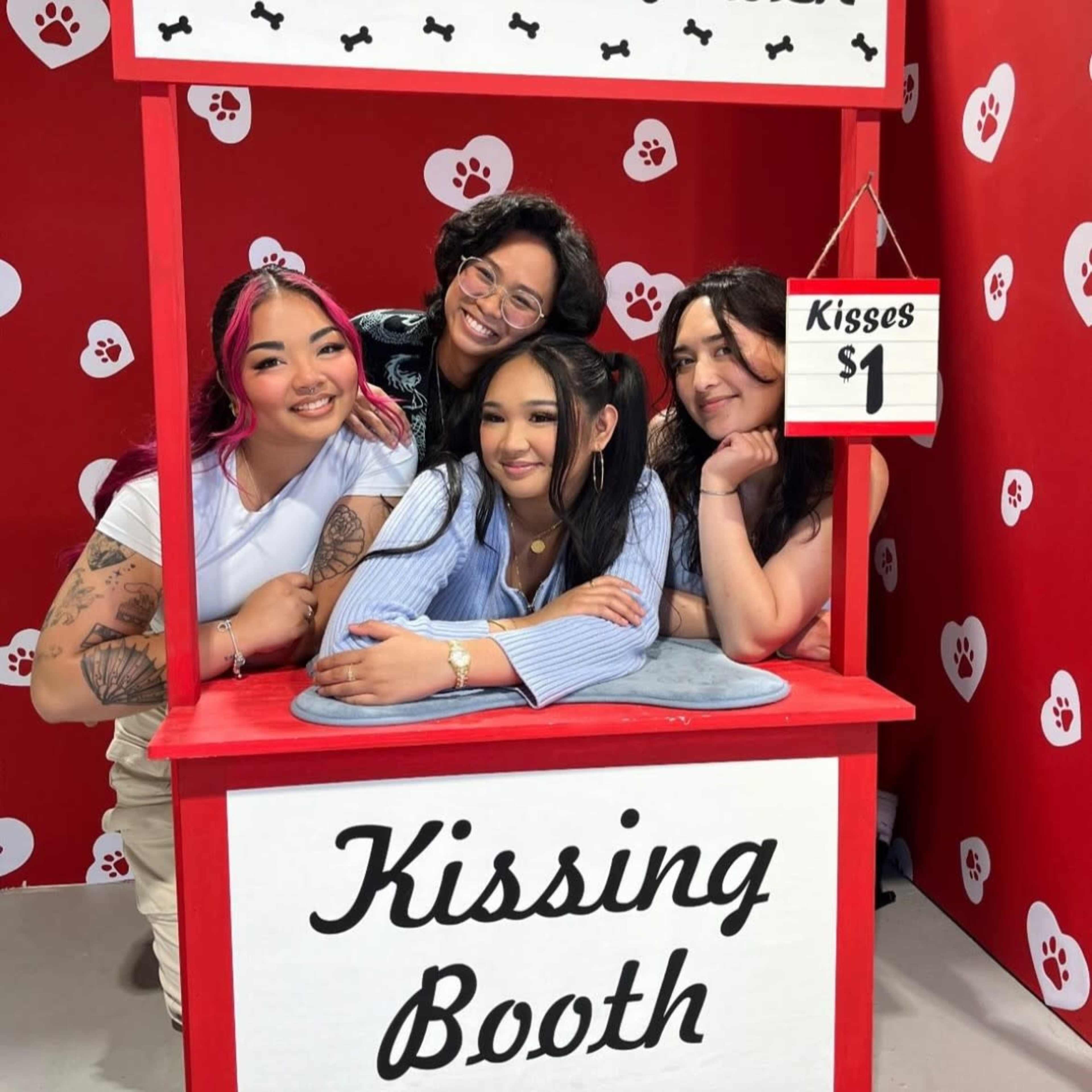 Four women pose together at a colorful kissing booth decorated with paw prints, with a sign indicating kisses are one dollar.
