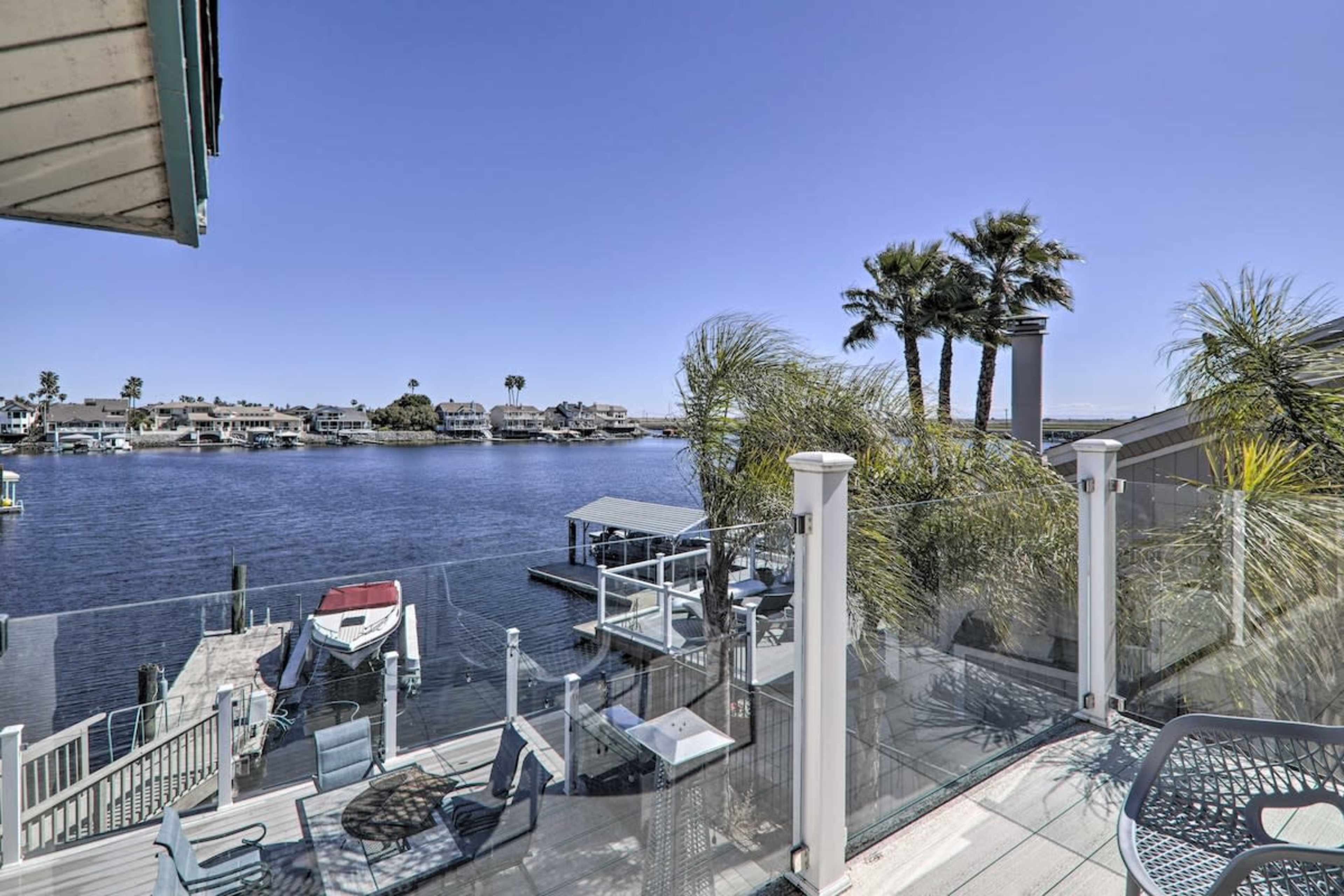 A waterfront terrace with glass railings, overlooking a calm bay lined with palm trees and houses on the opposite shore.