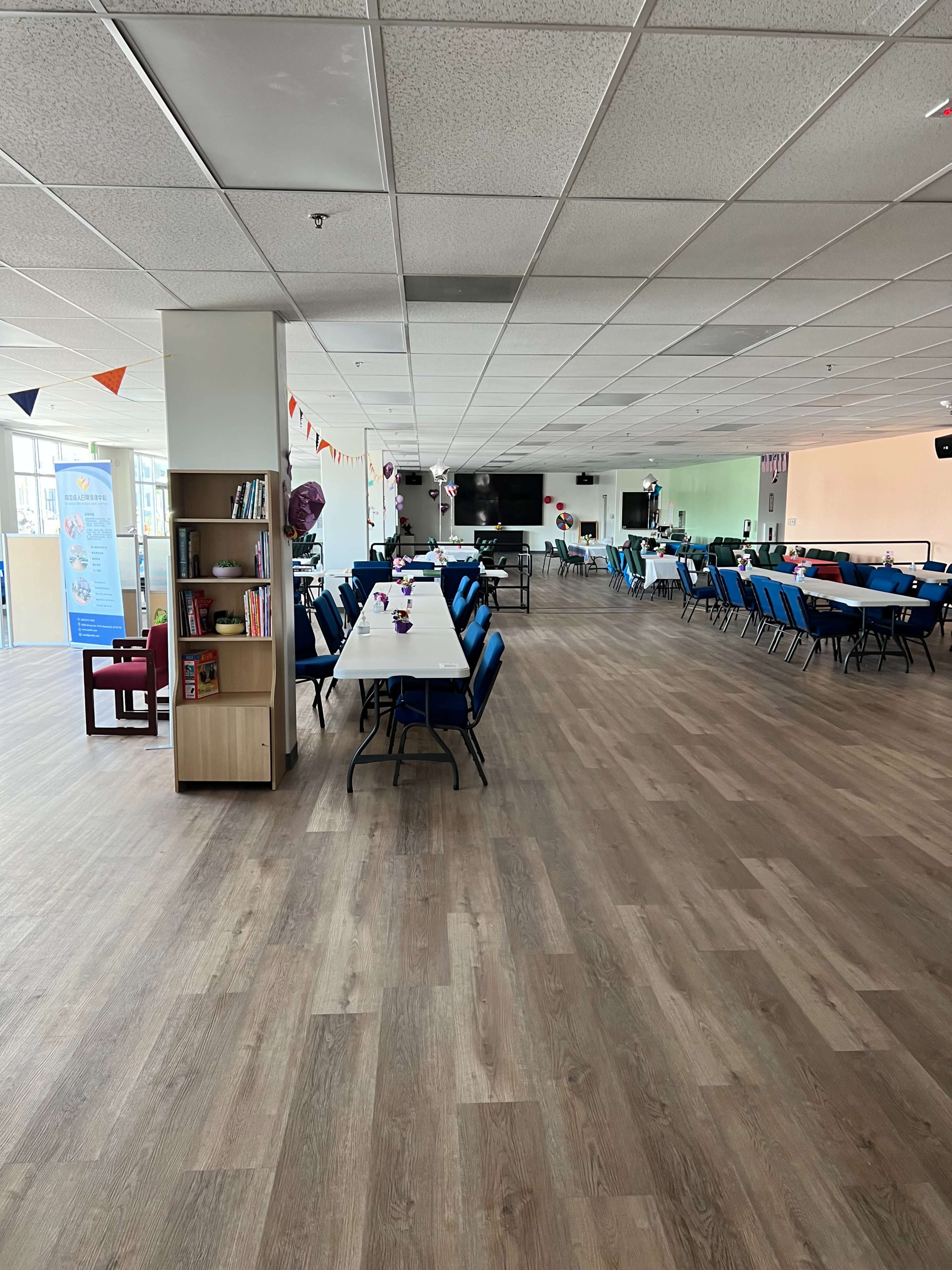 A spacious, well-lit room with blue chairs and tables arranged for a gathering, featuring wooden flooring and decorated with colorful banners.