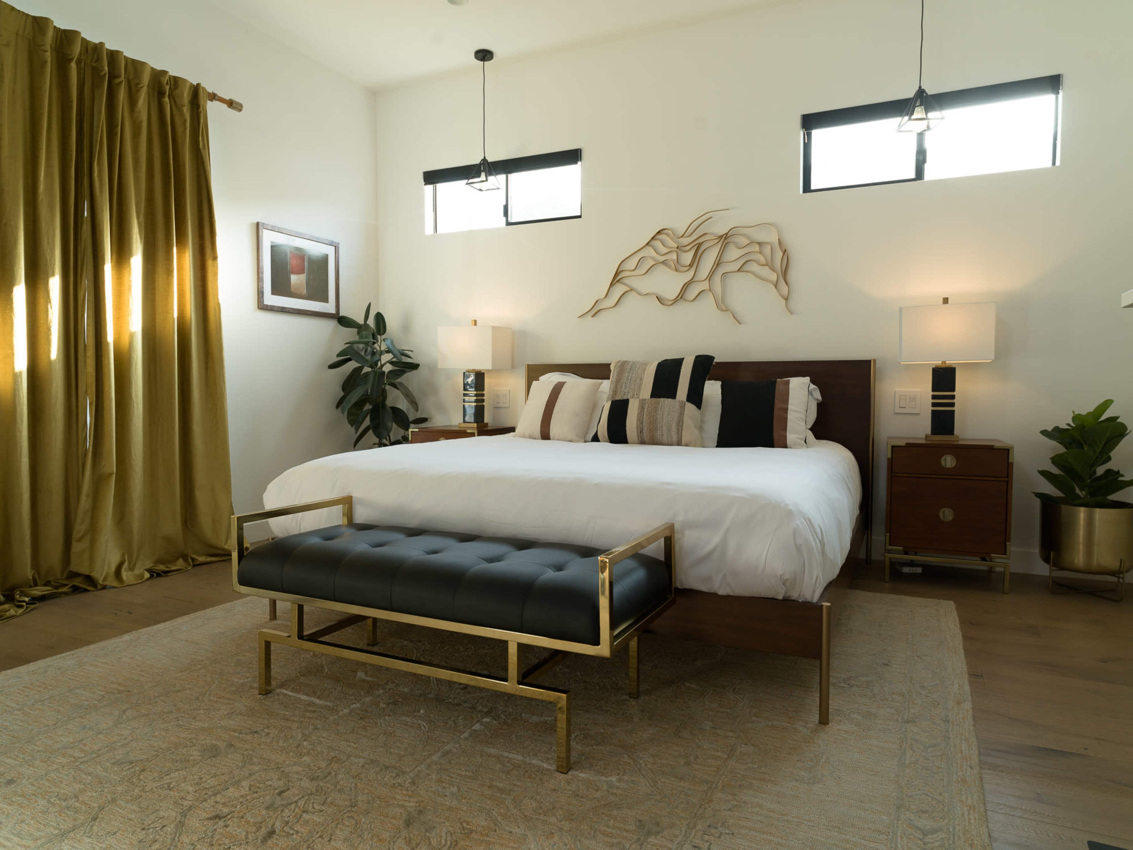 The image shows a modern bedroom featuring a large bed, two bedside lamps, a decorative wall piece, and a bench in front of the bed, with large windows allowing natural light to enter.