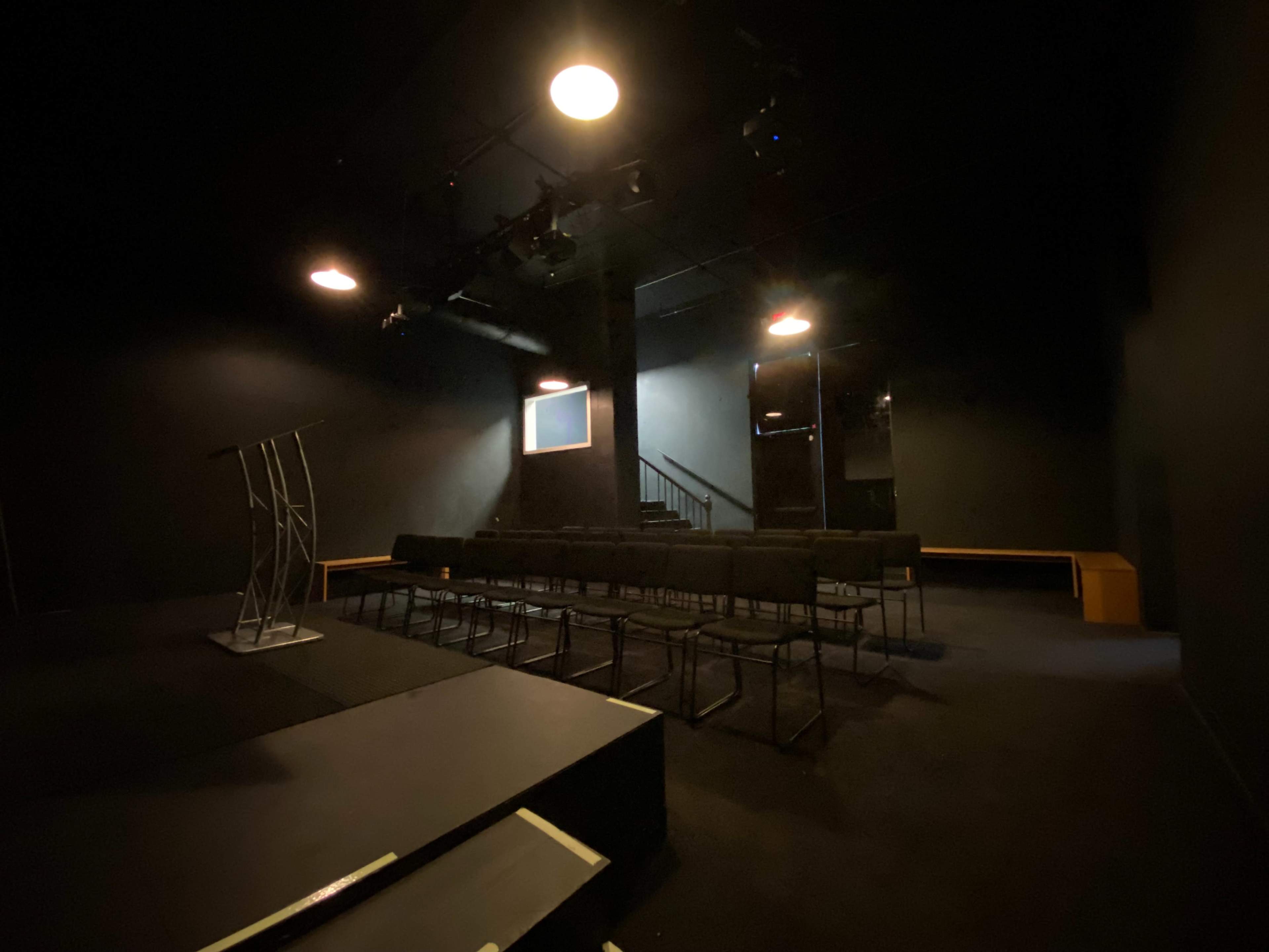 A dimly lit theater space with a podium, rows of chairs, and a staircase in the background.