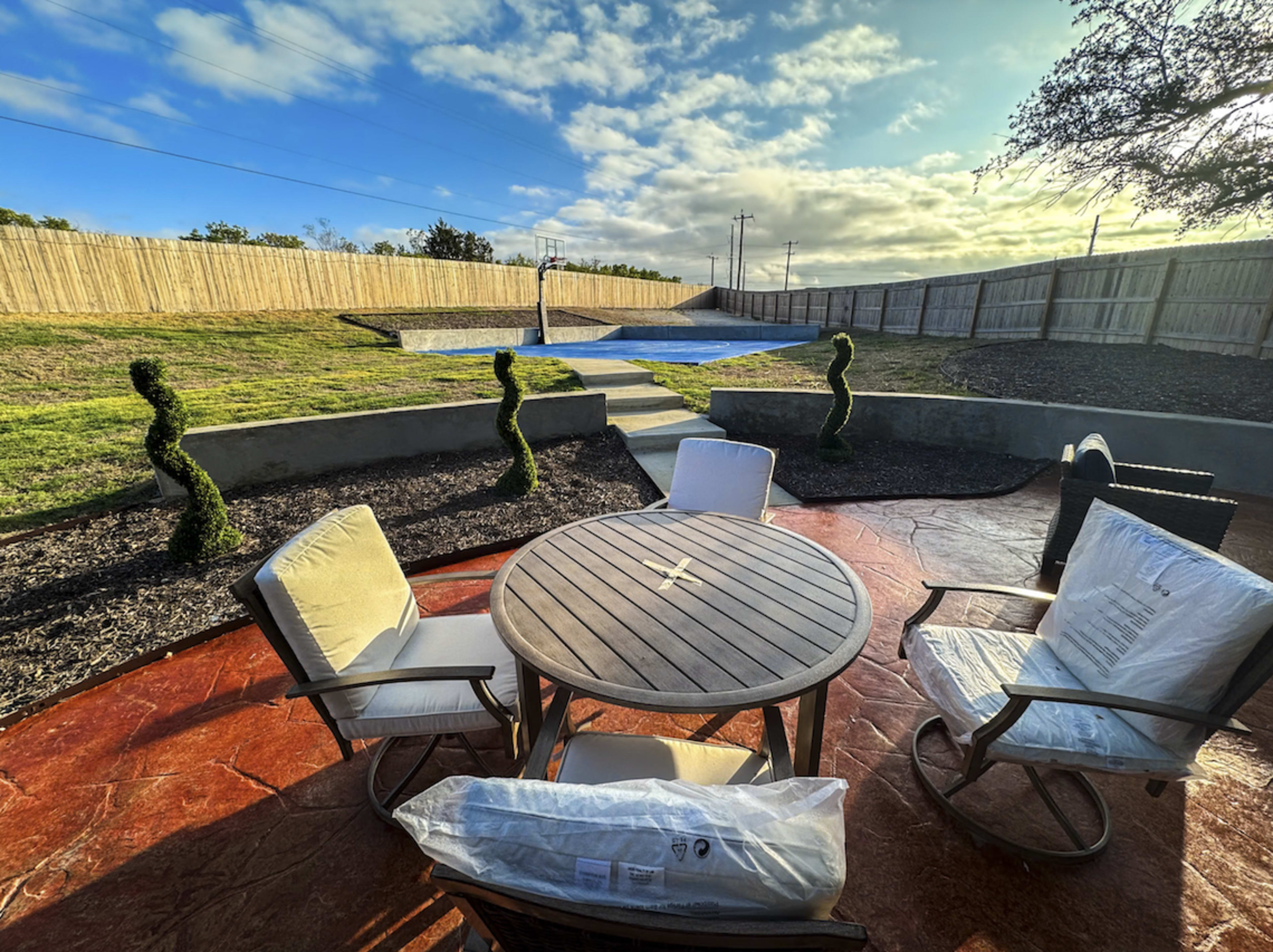 The image shows a landscaped backyard with a circular wooden table surrounded by four chairs, topped with hedges and a covered pool in the background.