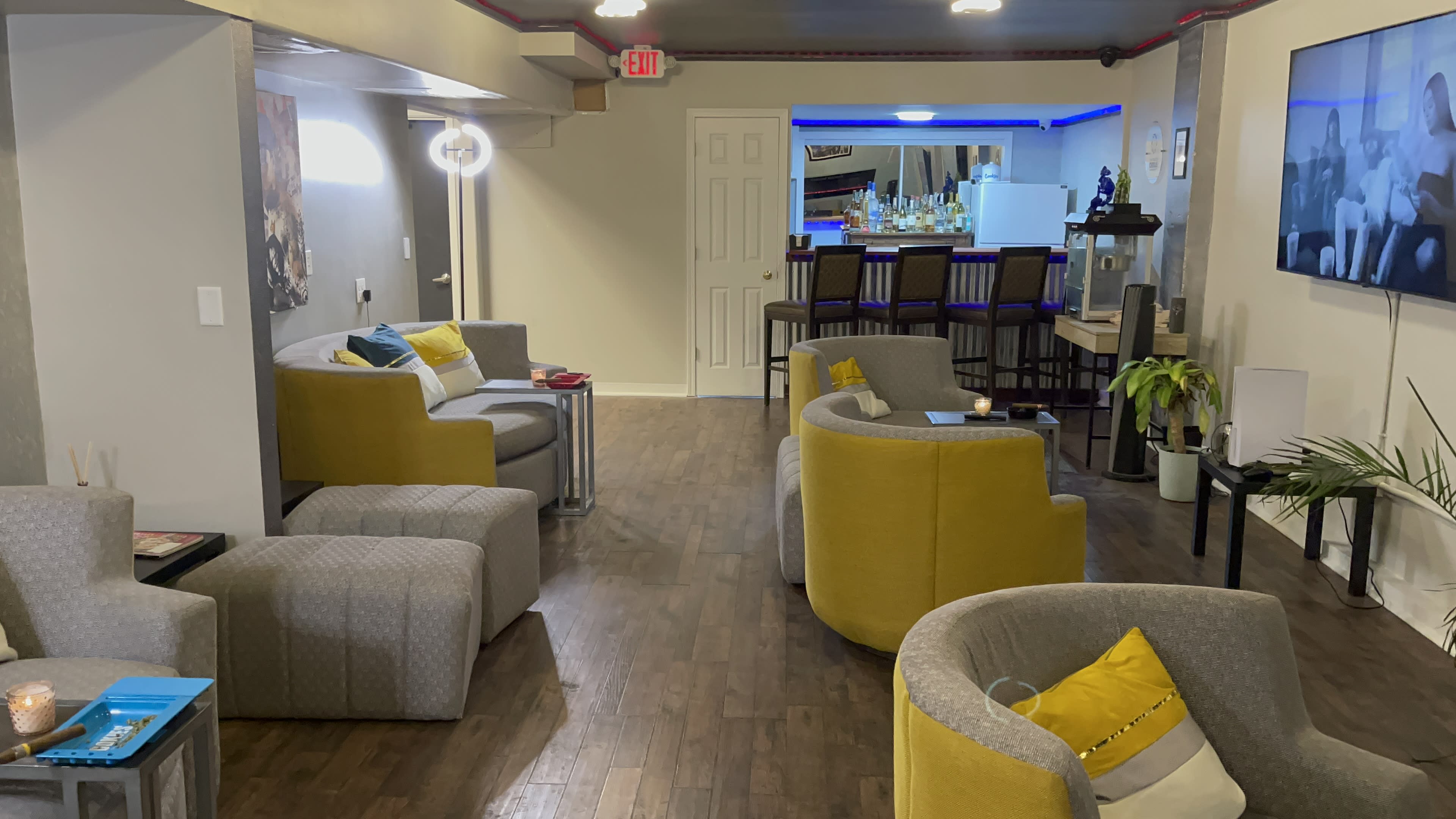 A modern lounge area with gray and yellow seating arrangements, wooden flooring, and a bar in the background with blue lighting.