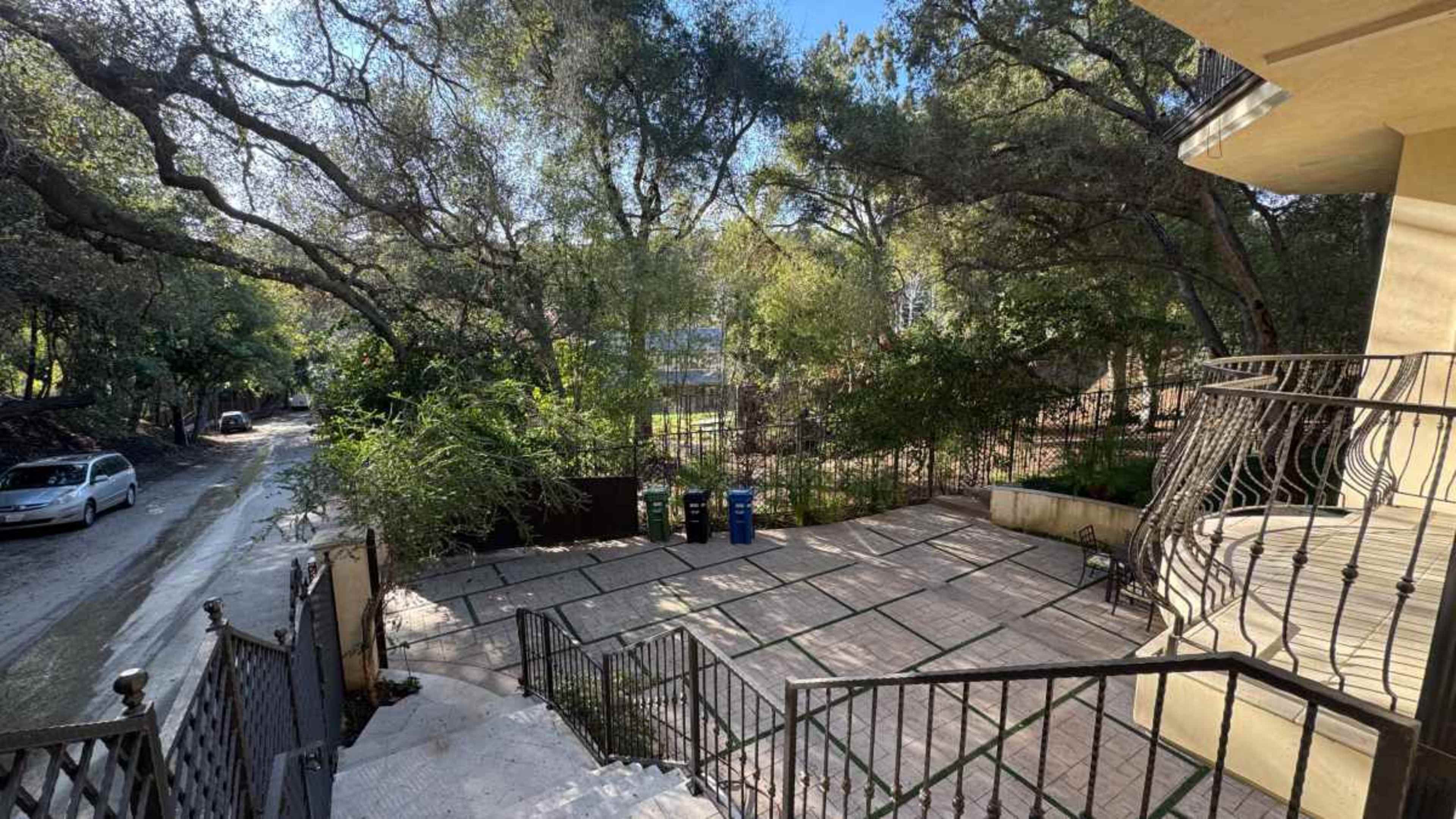 The image shows a pathway leading down from a house, surrounded by trees, with a view of a street and several trash bins.