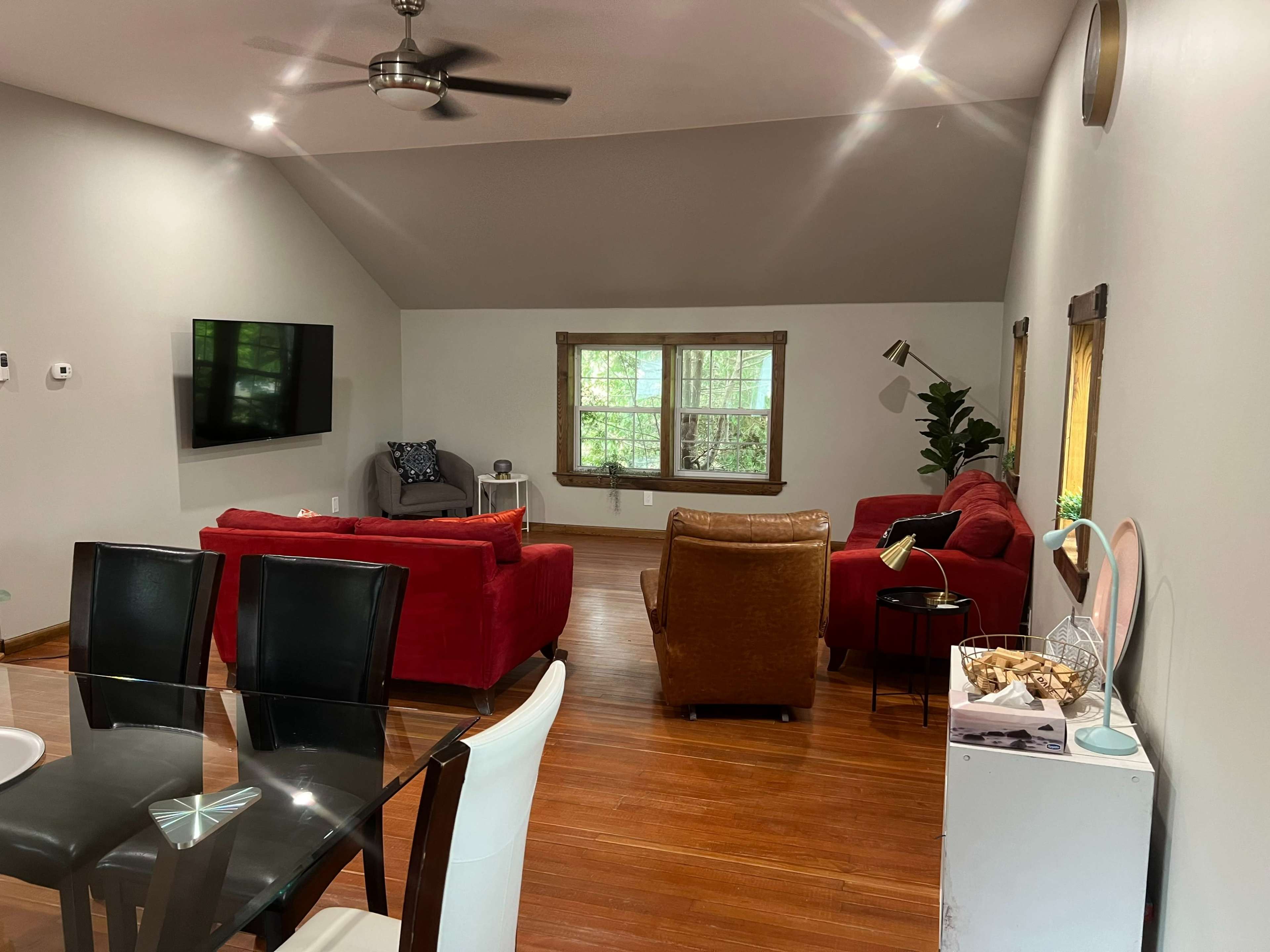 A living room features red sofas, a brown armchair, a dining table, and large windows with greenery outside.