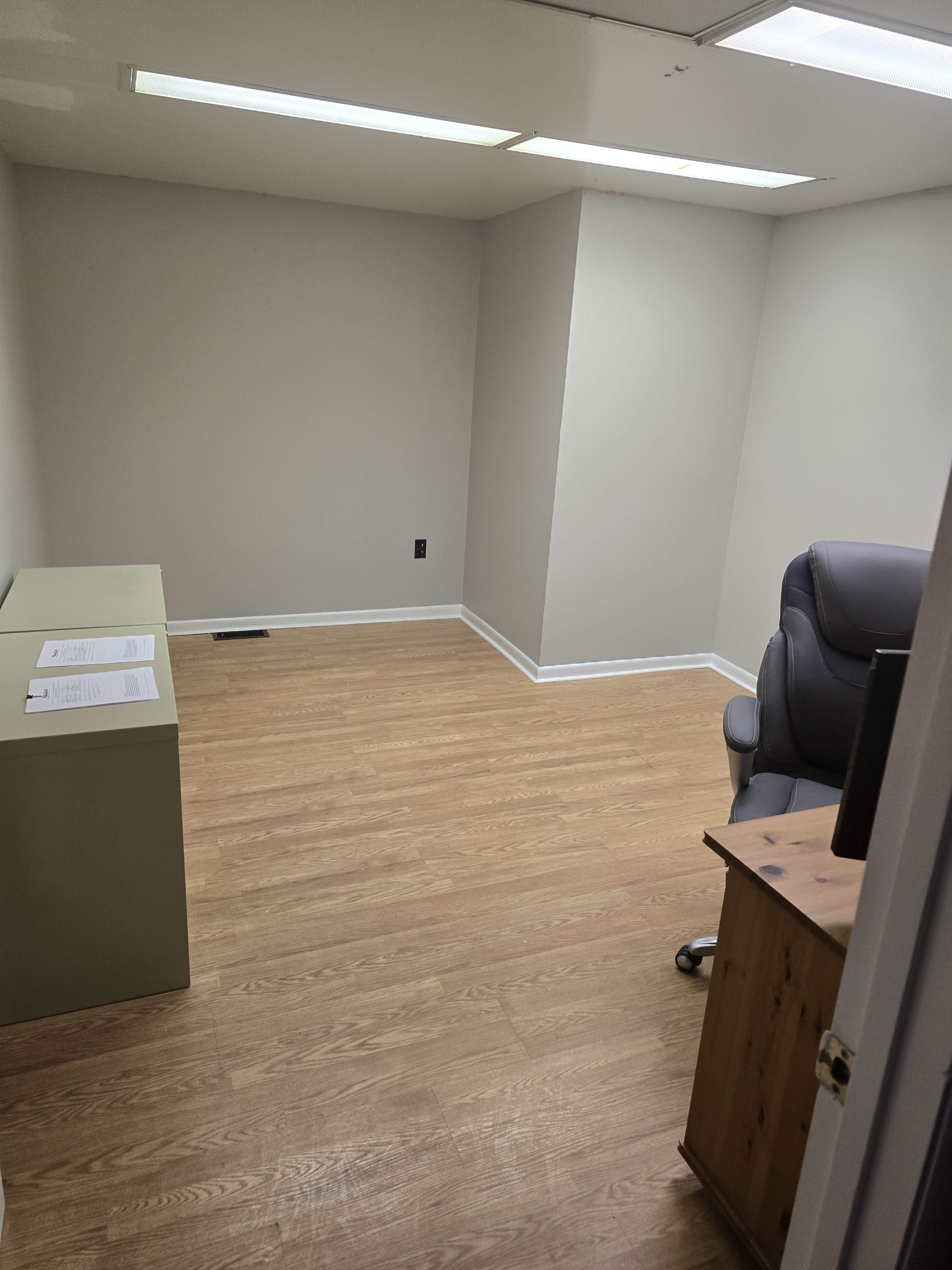 The image shows a sparsely furnished office with light-colored walls, wooden flooring, a desk, a gray office chair, and some papers on a countertop.