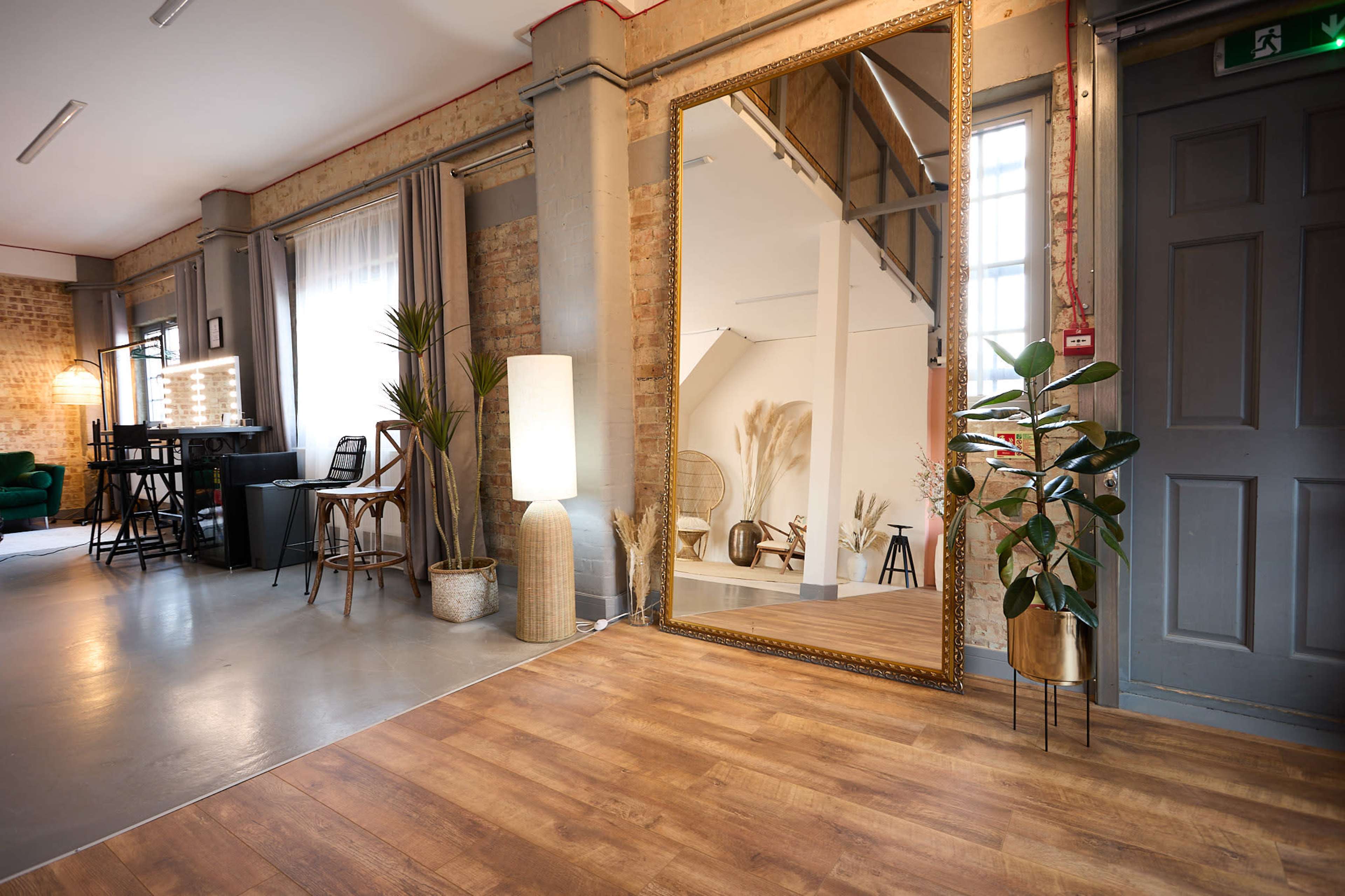 The image shows a spacious interior with hardwood flooring, featuring a large decorative mirror, various plants, and furniture like chairs and a lamp against exposed brick walls.