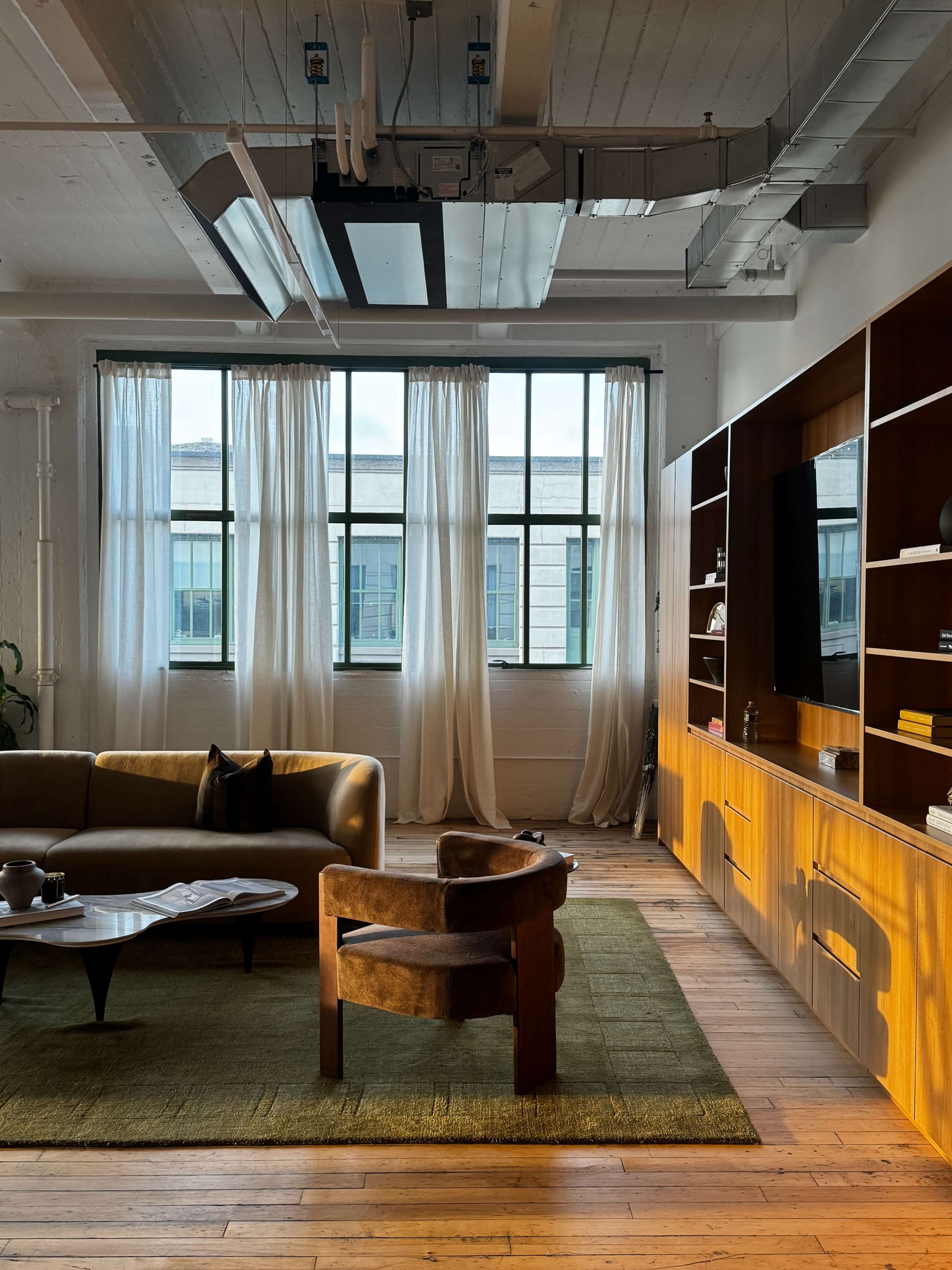 A bright and airy living room features large windows with sheer curtains, modern furniture including a brown sofa and chair, and wooden shelving against the wall.