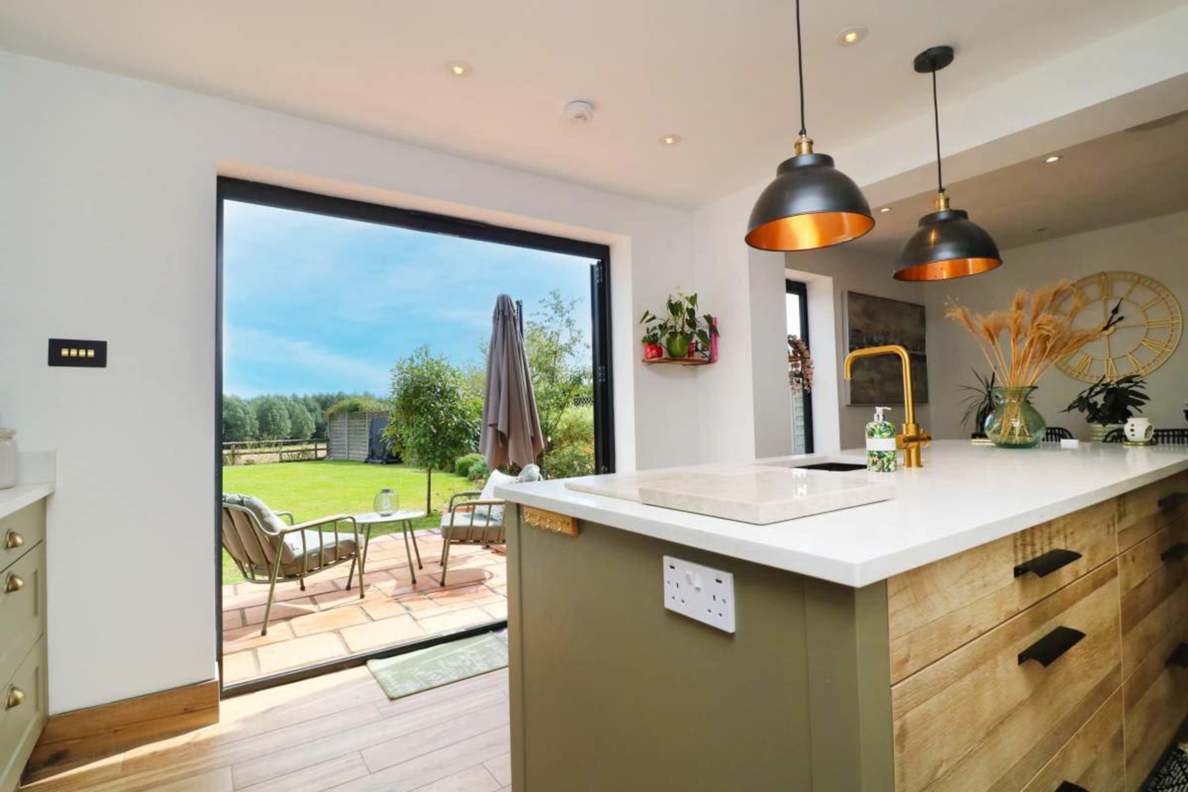 A modern kitchen features a large island with a sink and pendant lights, opening up to a patio overlooking a green garden.