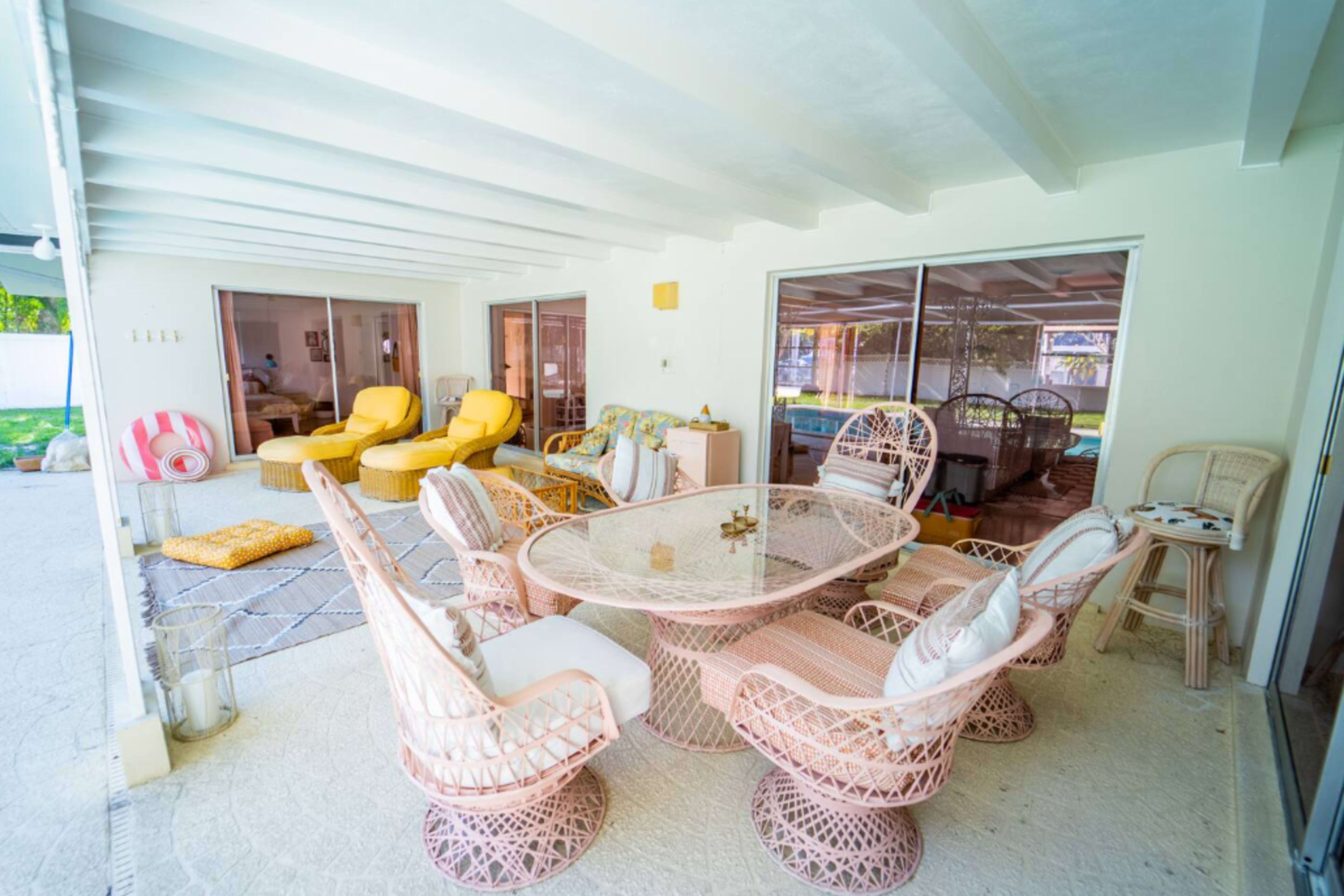 The image features a covered patio area furnished with a round glass table surrounded by wicker chairs, yellow lounge chairs, and a view of a pool through large sliding glass doors.