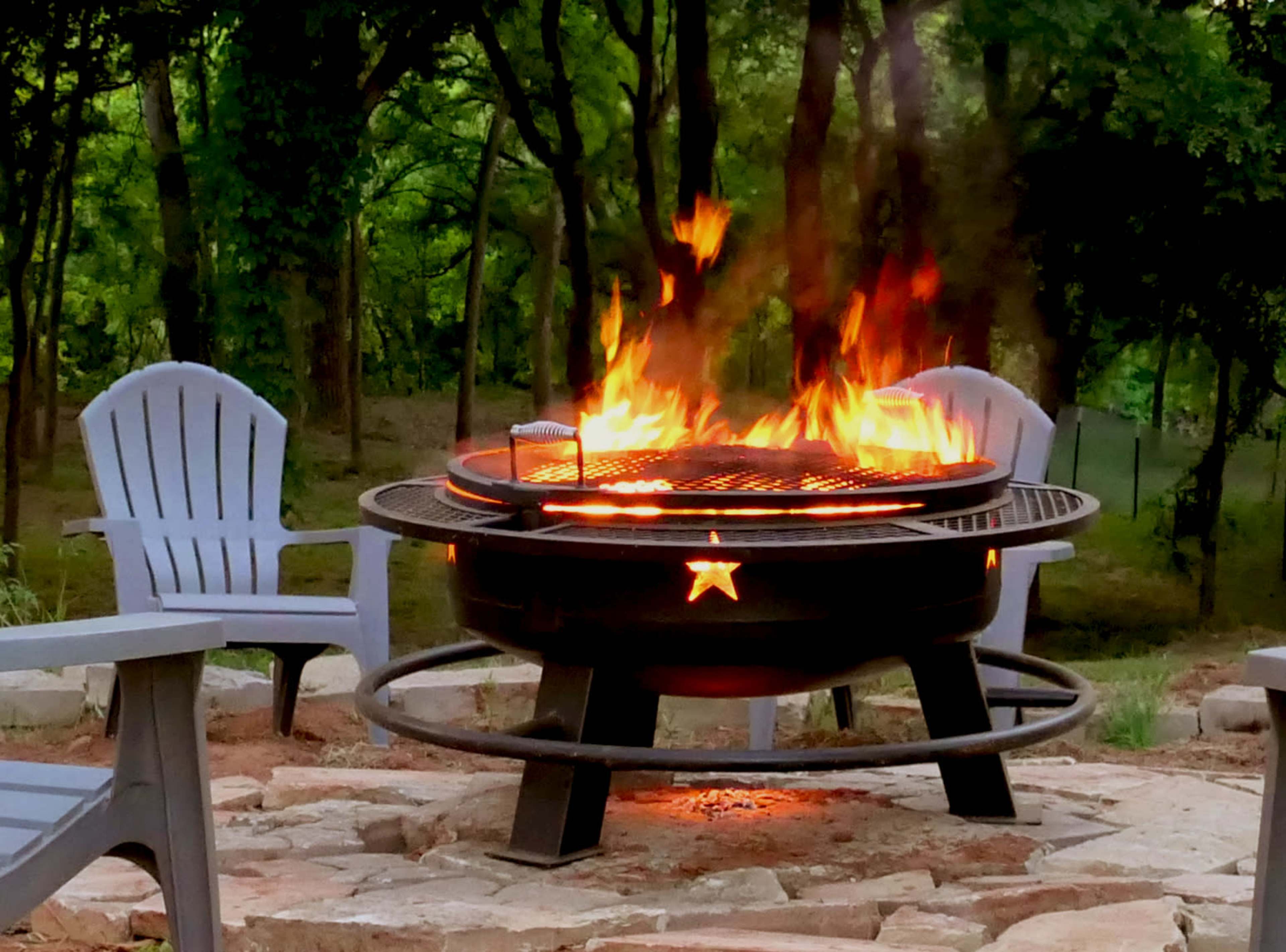 A fire pit surrounded by chairs emits bright flames in a wooded area.
