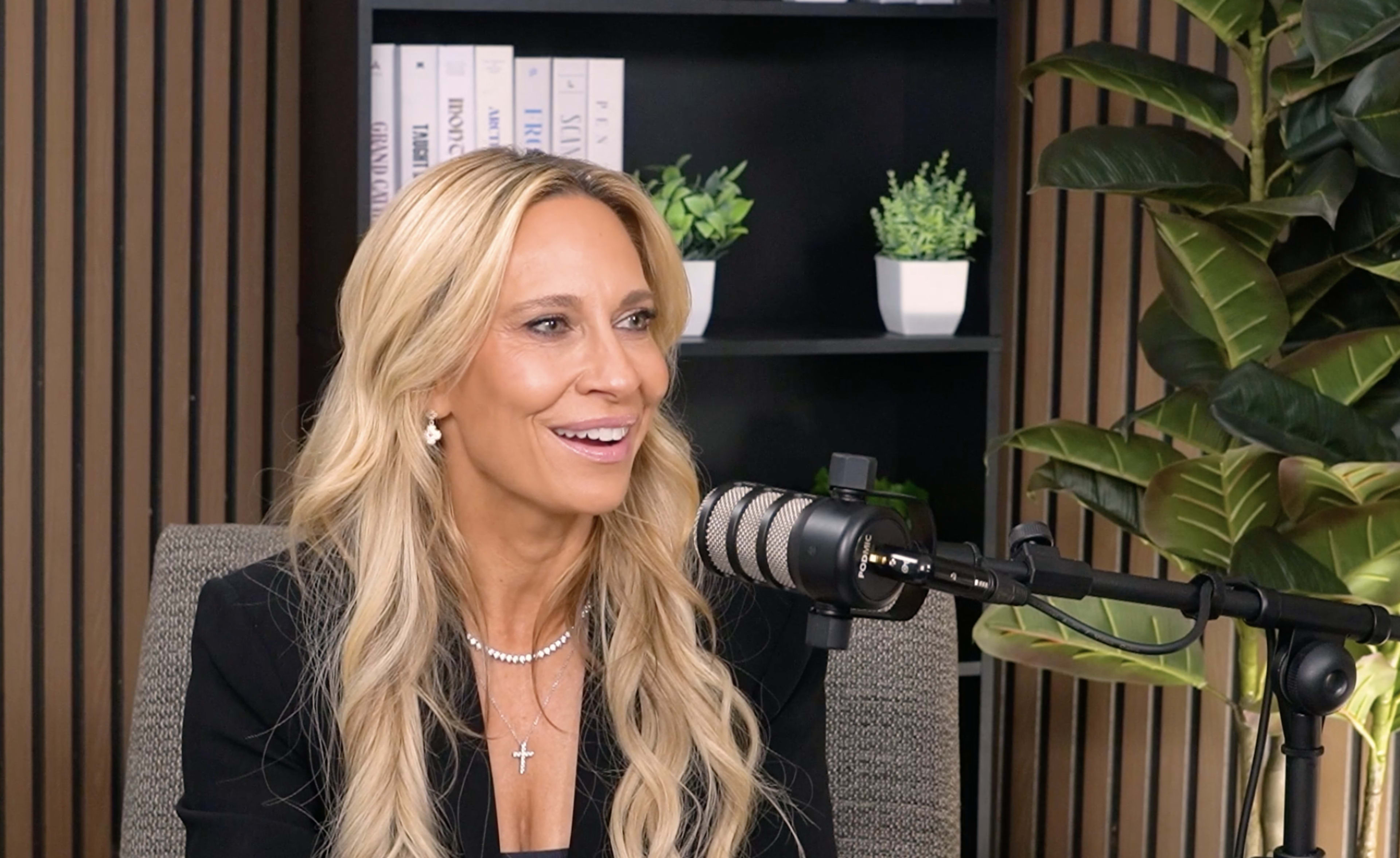 A woman with long, blonde hair is smiling and speaking into a microphone while seated in front of a bookshelf and potted plants.