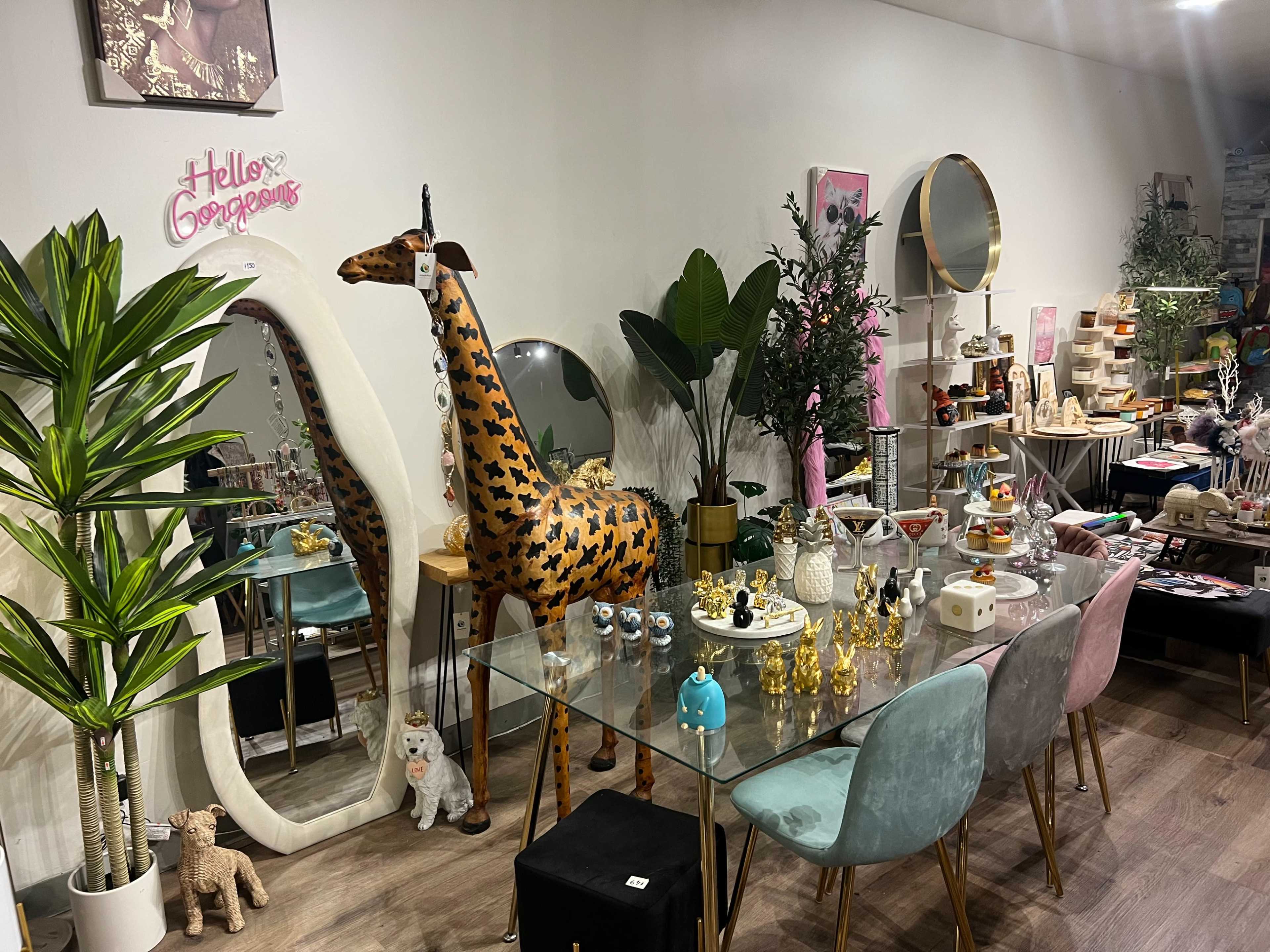 The image shows a boutique interior featuring a glass table adorned with various decorative items and a tall, painted giraffe statue surrounded by plants and colorful seating.