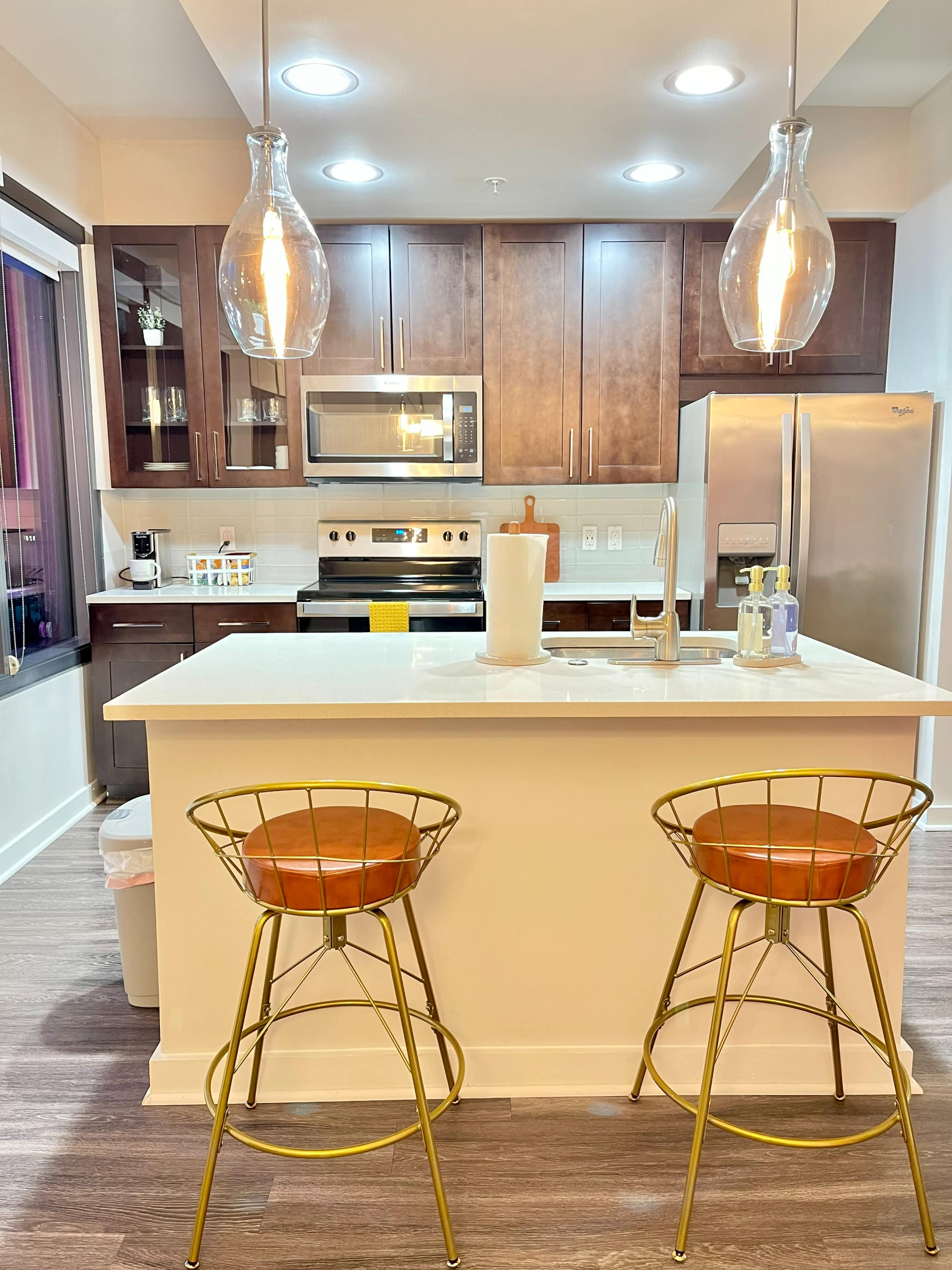 A modern kitchen features dark wood cabinetry, stainless steel appliances, and two gold wire bar stools with brown leather seats at a white island.
