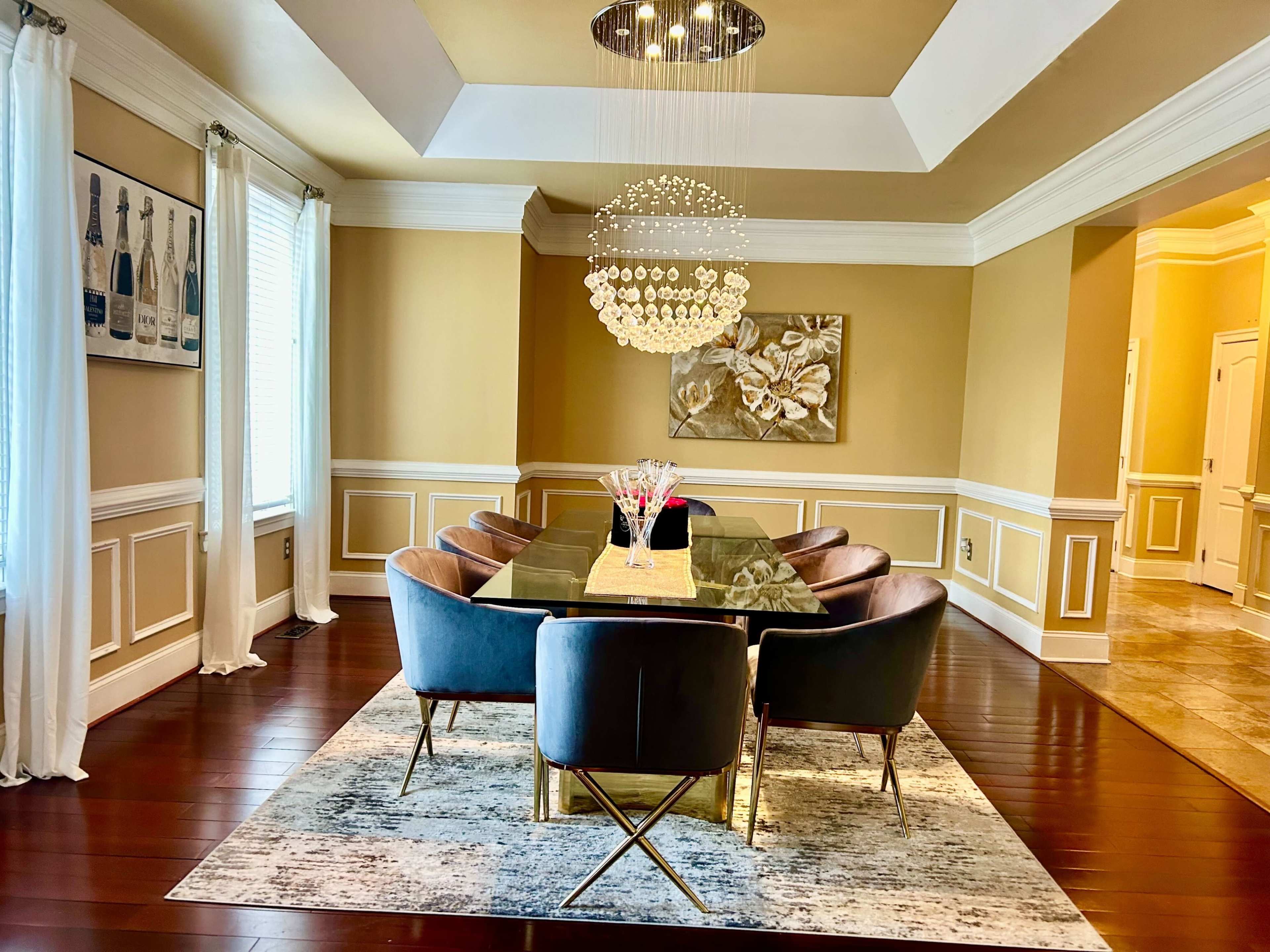 A modern dining room featuring a large glass table surrounded by eight upholstered chairs, with a chandelier overhead and decorative wall art.