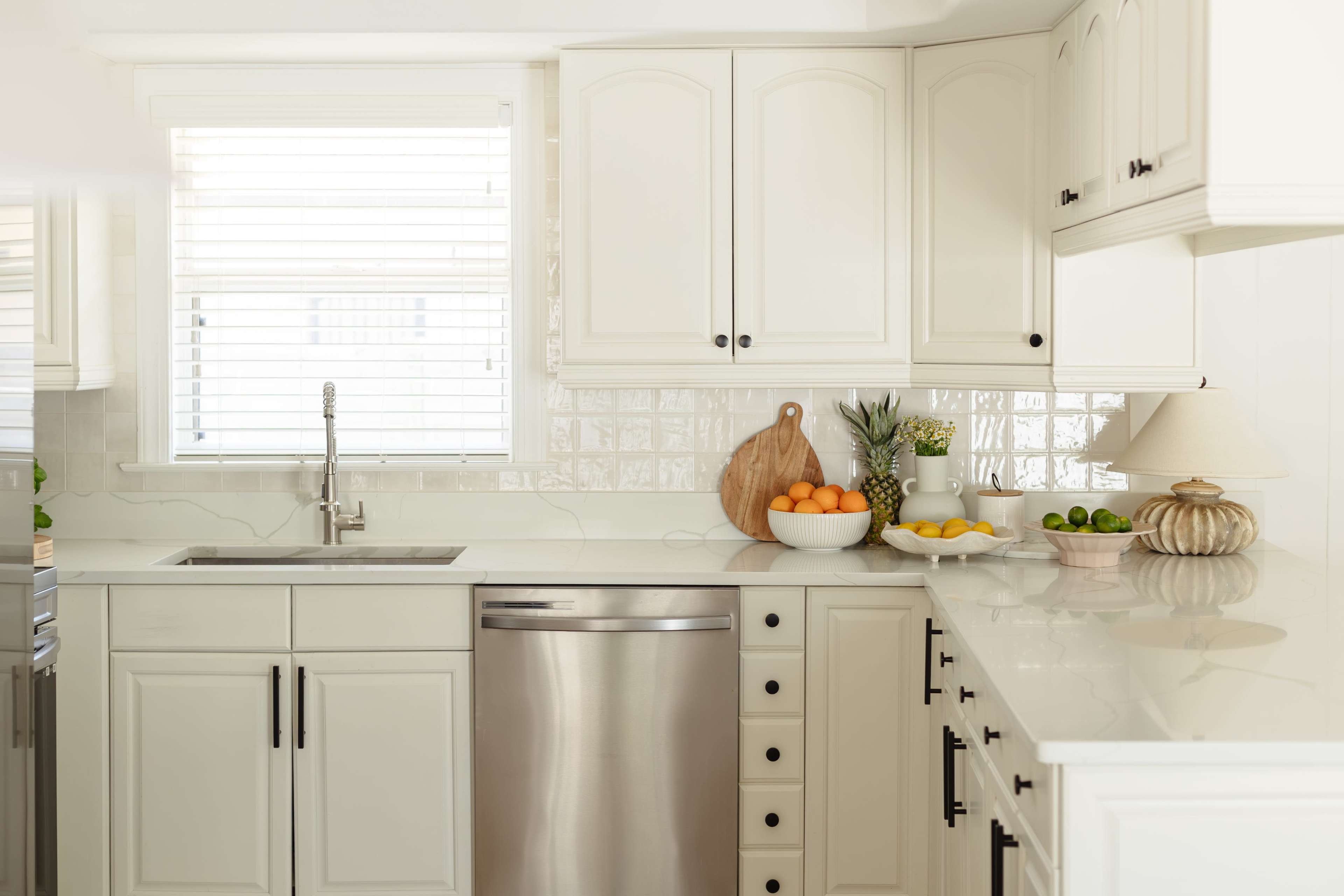 The image shows a modern kitchen with white cabinetry, a stainless steel dishwasher, and a countertop adorned with a variety of fresh fruits.