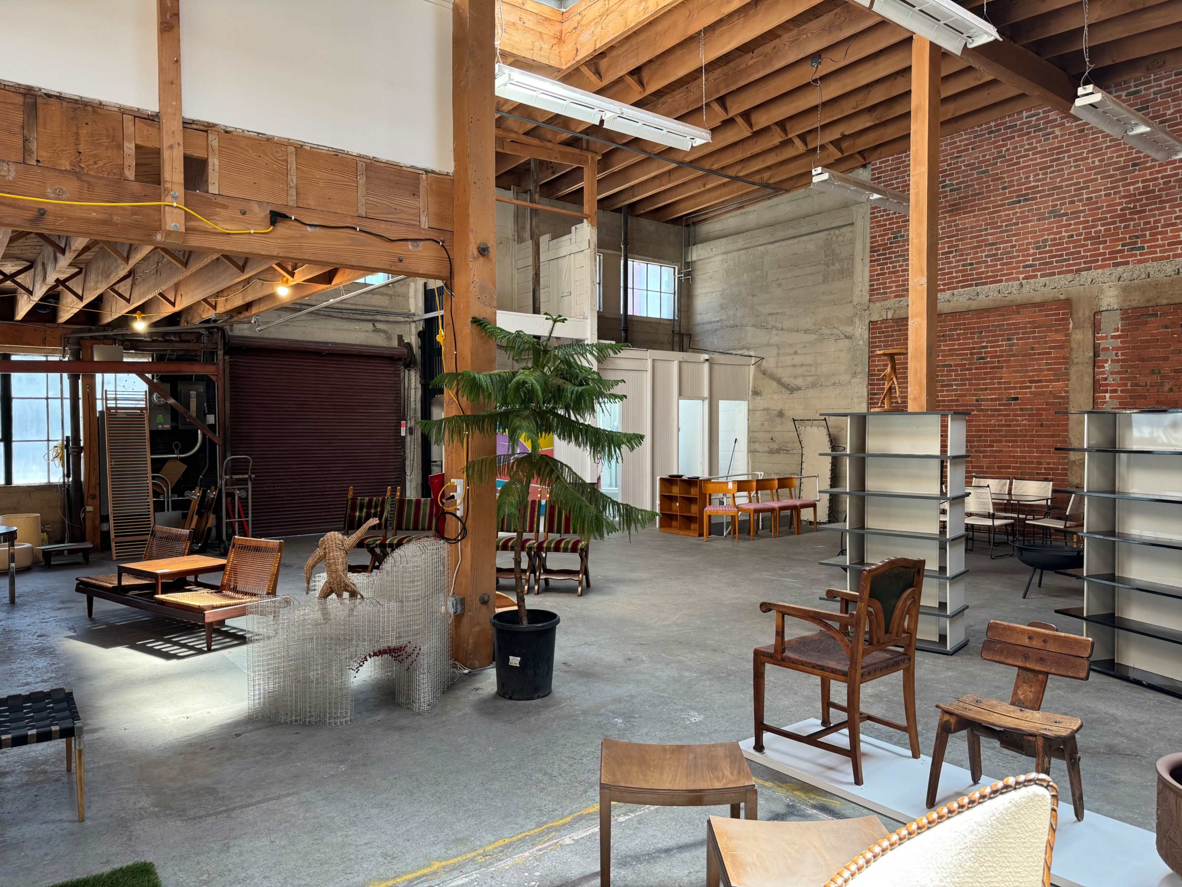 An industrial-style interior space filled with various types of furniture, including chairs, tables, and shelving units, under a wooden roof with exposed beams.