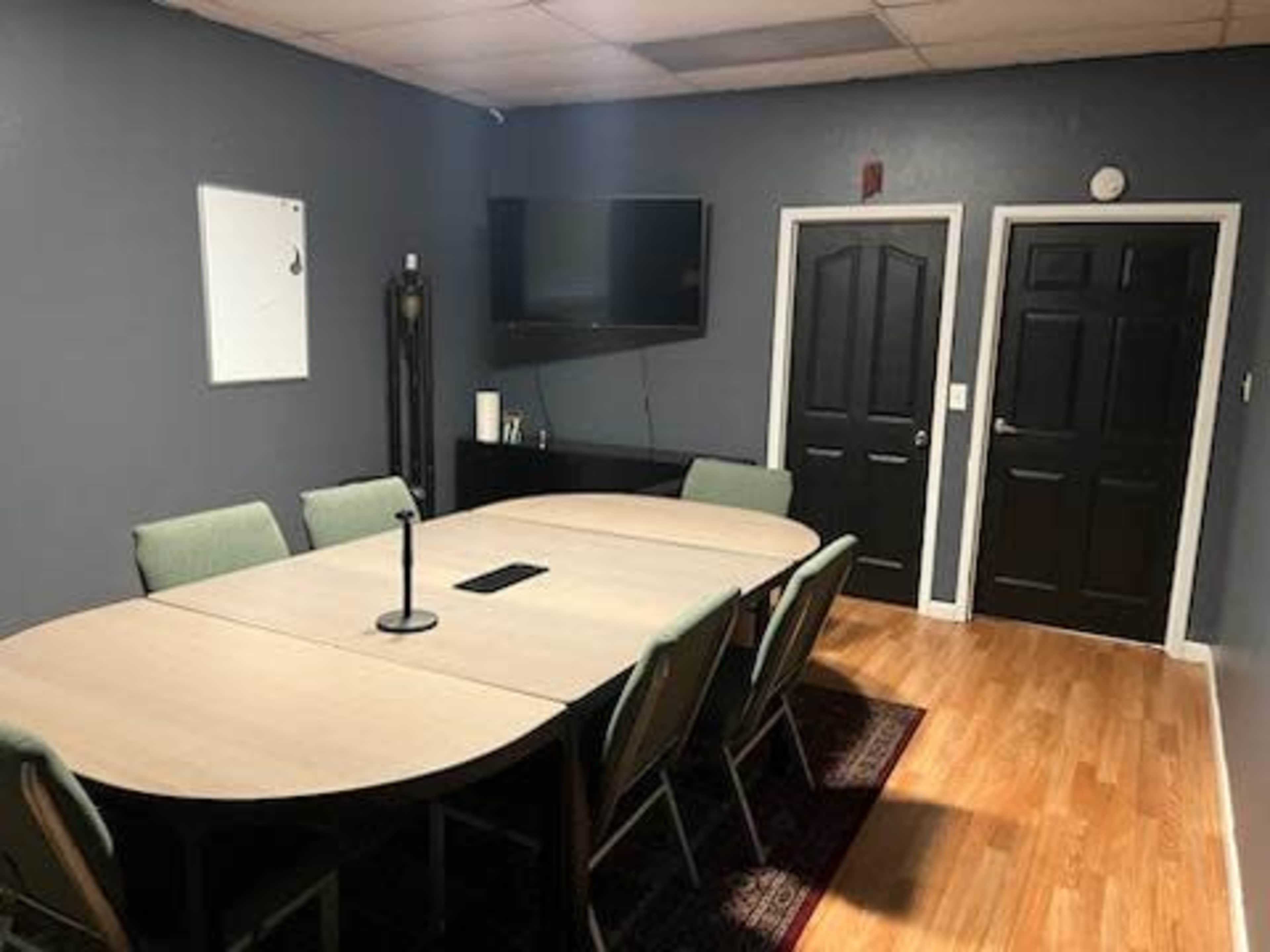 A conference room with a large oval table surrounded by chairs, a television mounted on the wall, and two closed doors.