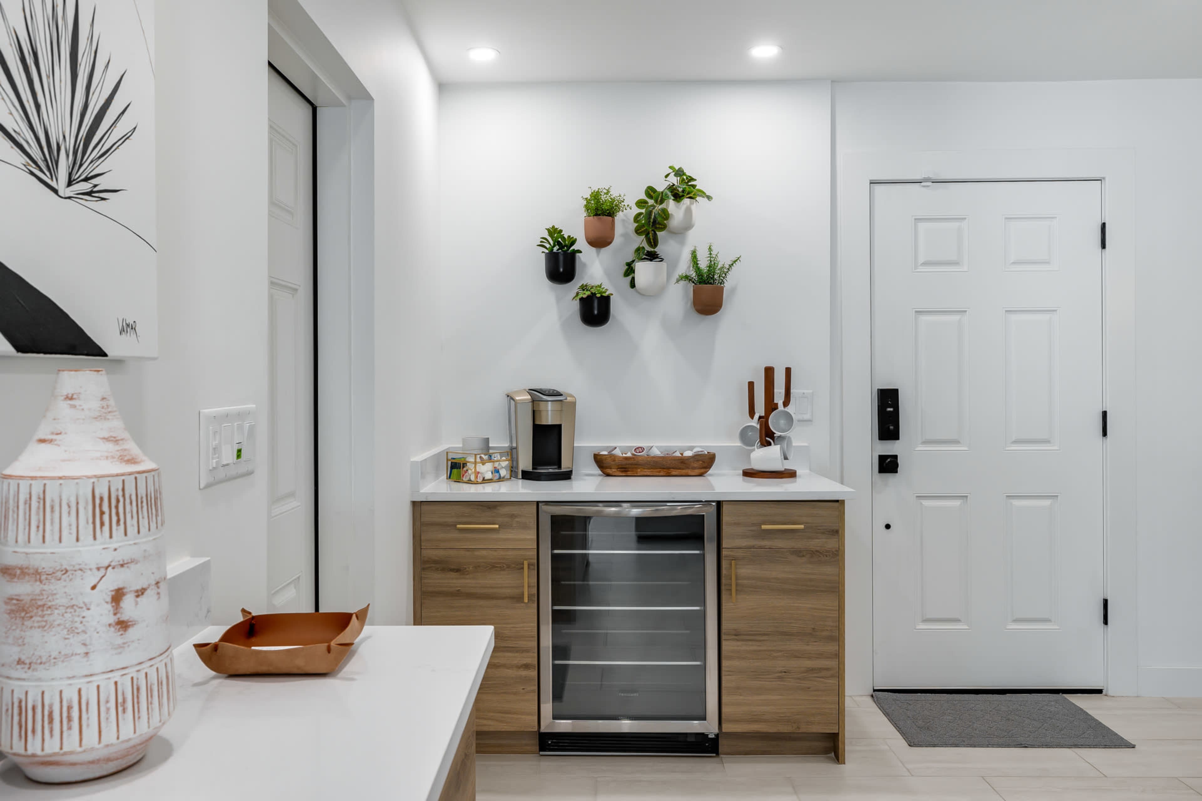 A modern kitchen area features a compact beverage cooler, a coffee maker, wall-mounted planters with plants, and a decorative bowl on a countertop.