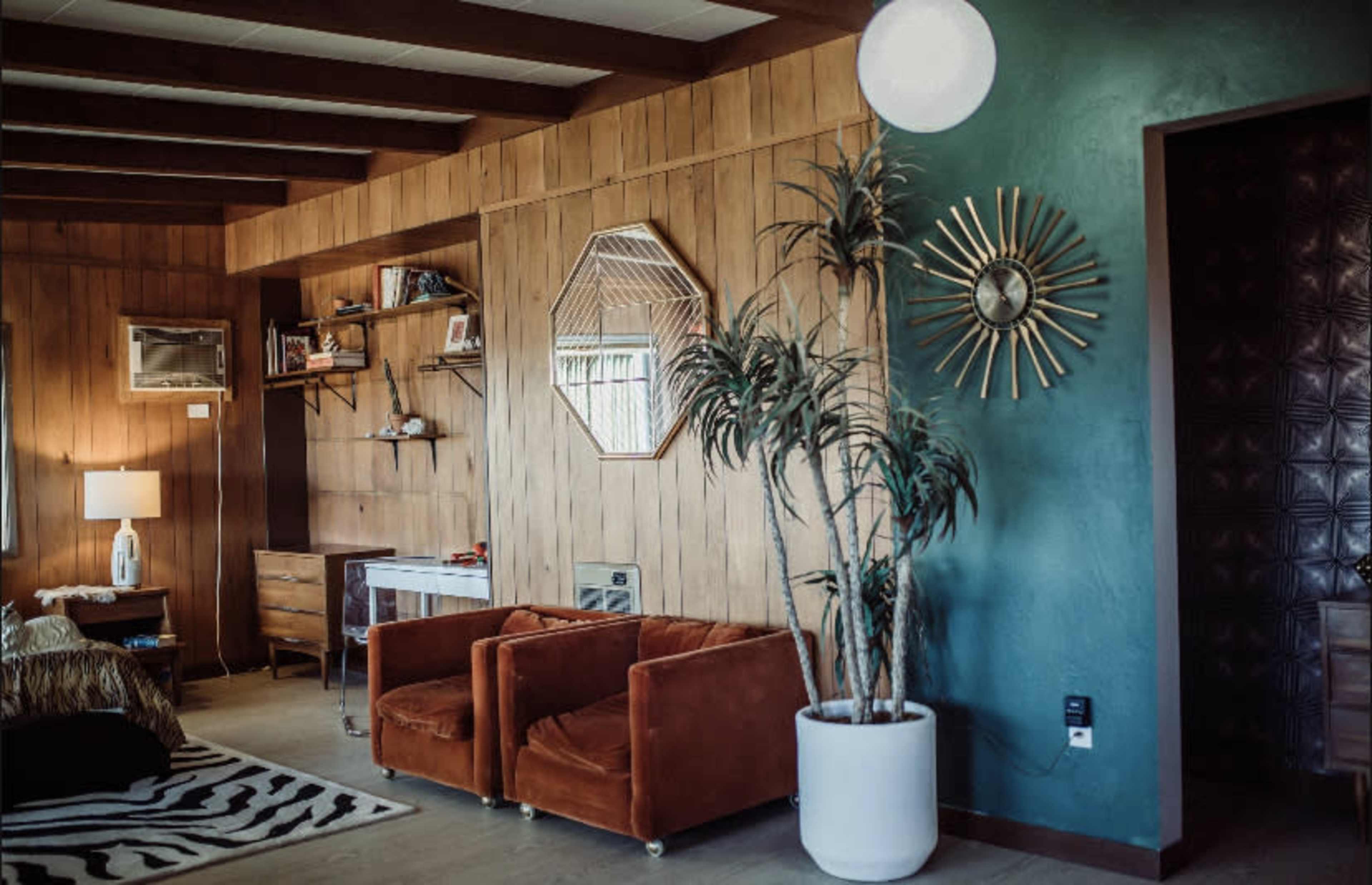 The image showcases a living room with wooden paneling, two velvet orange chairs, a potted plant, and a wall clock.