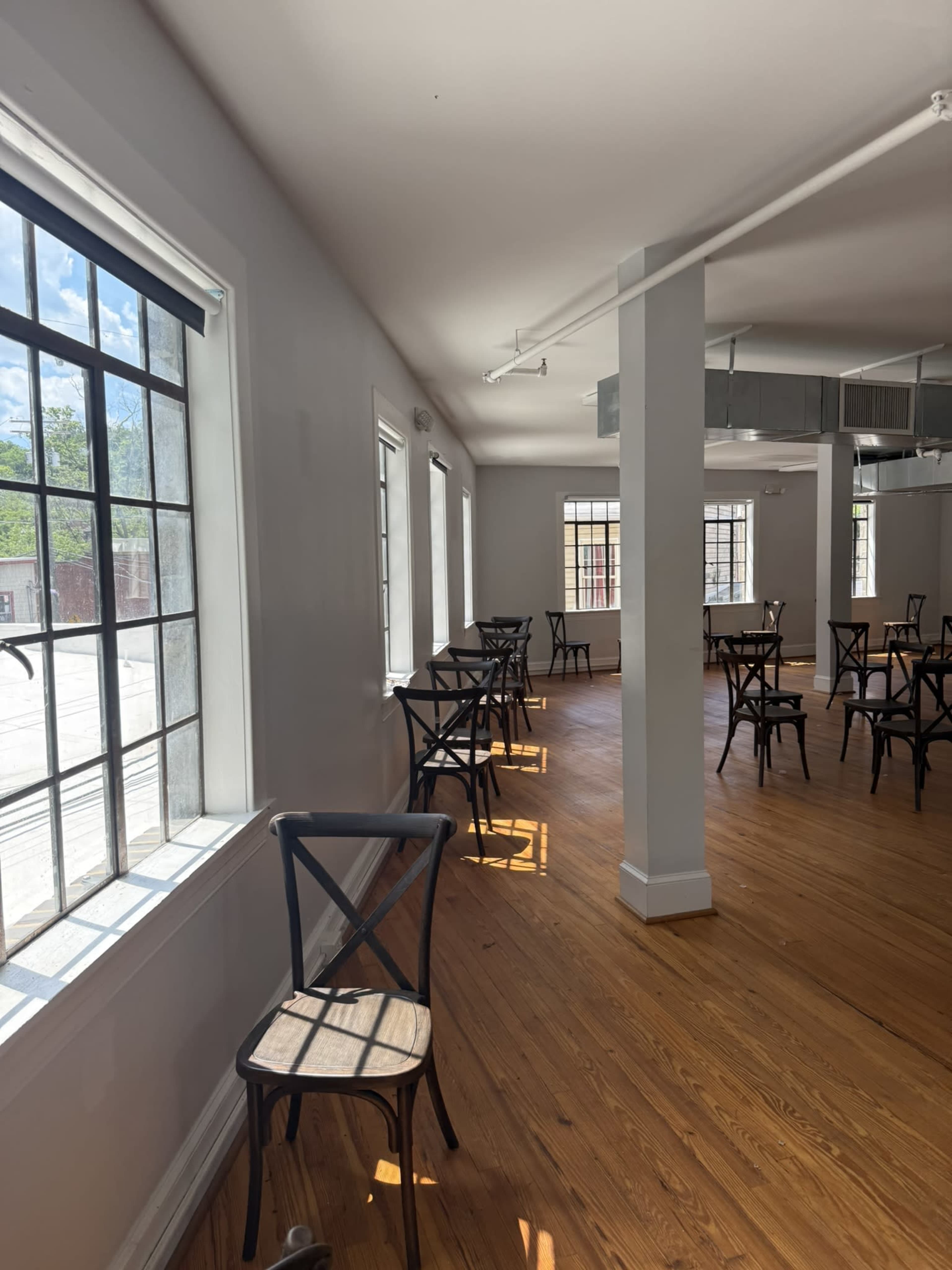 The image shows a spacious room with wooden floors, featuring several black chairs and large windows allowing natural light to enter.