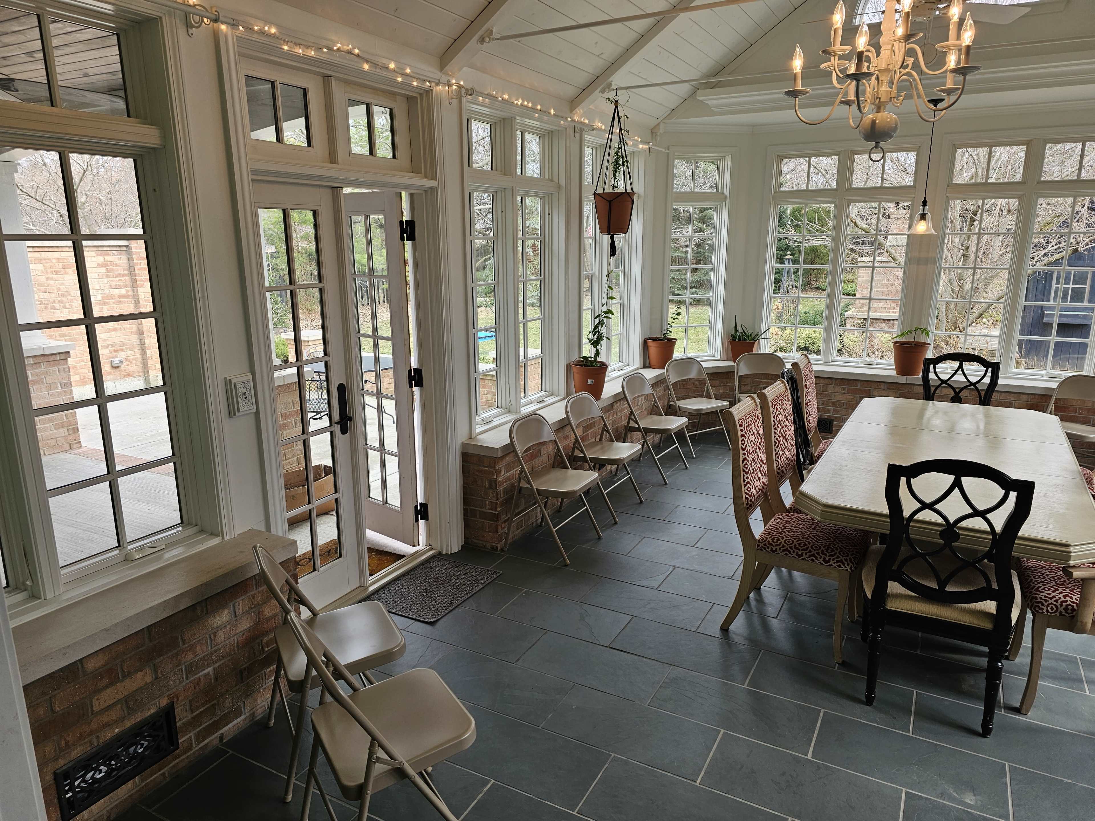The image shows a sunlit room with large windows, featuring a long dining table surrounded by chairs and a door leading outside.