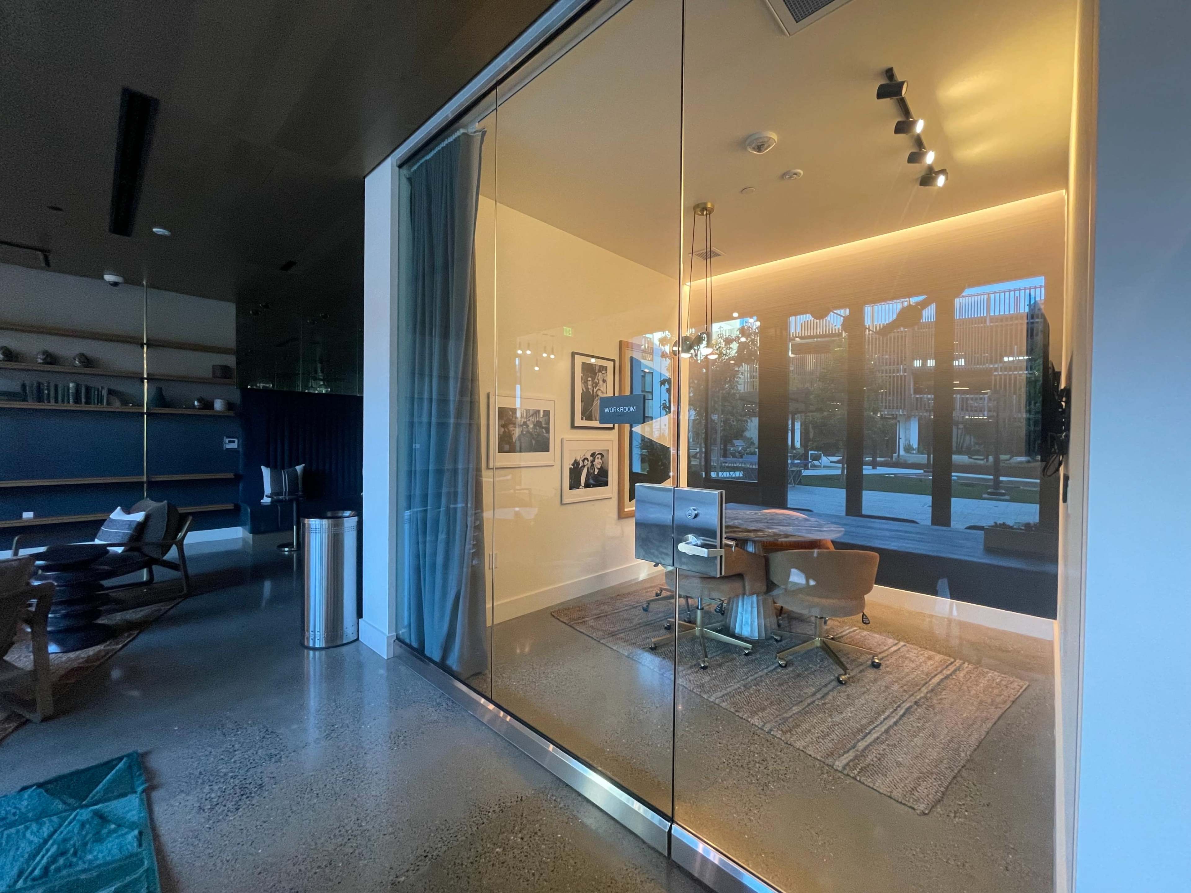 A modern office space with a glass wall, featuring a desk and chair, illuminated by warm lighting, and framed photographs on the wall.
