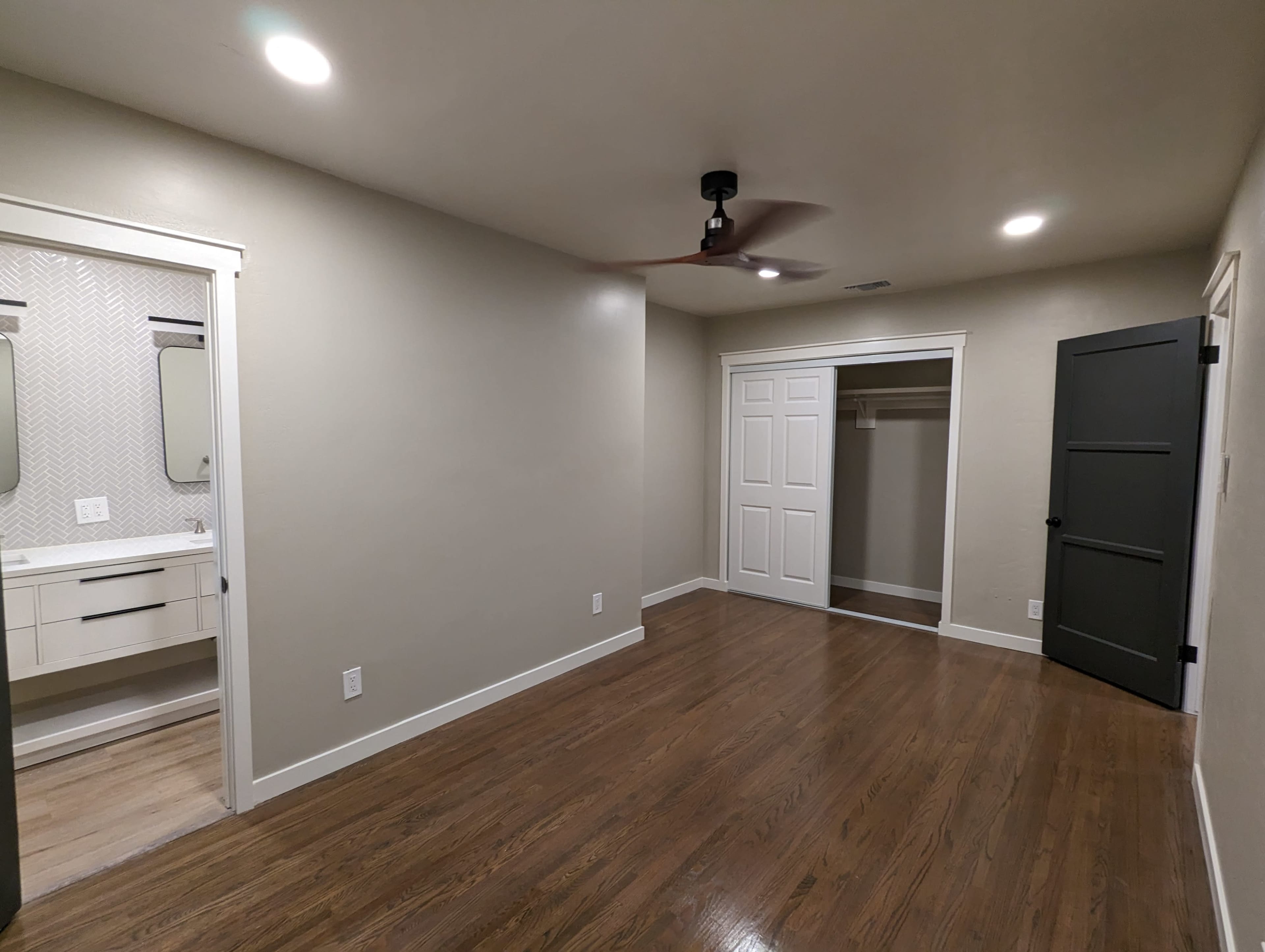 The image shows a well-lit room with hardwood floors, featuring a closet with a sliding door and a bathroom area with two mirrors and a vanity.