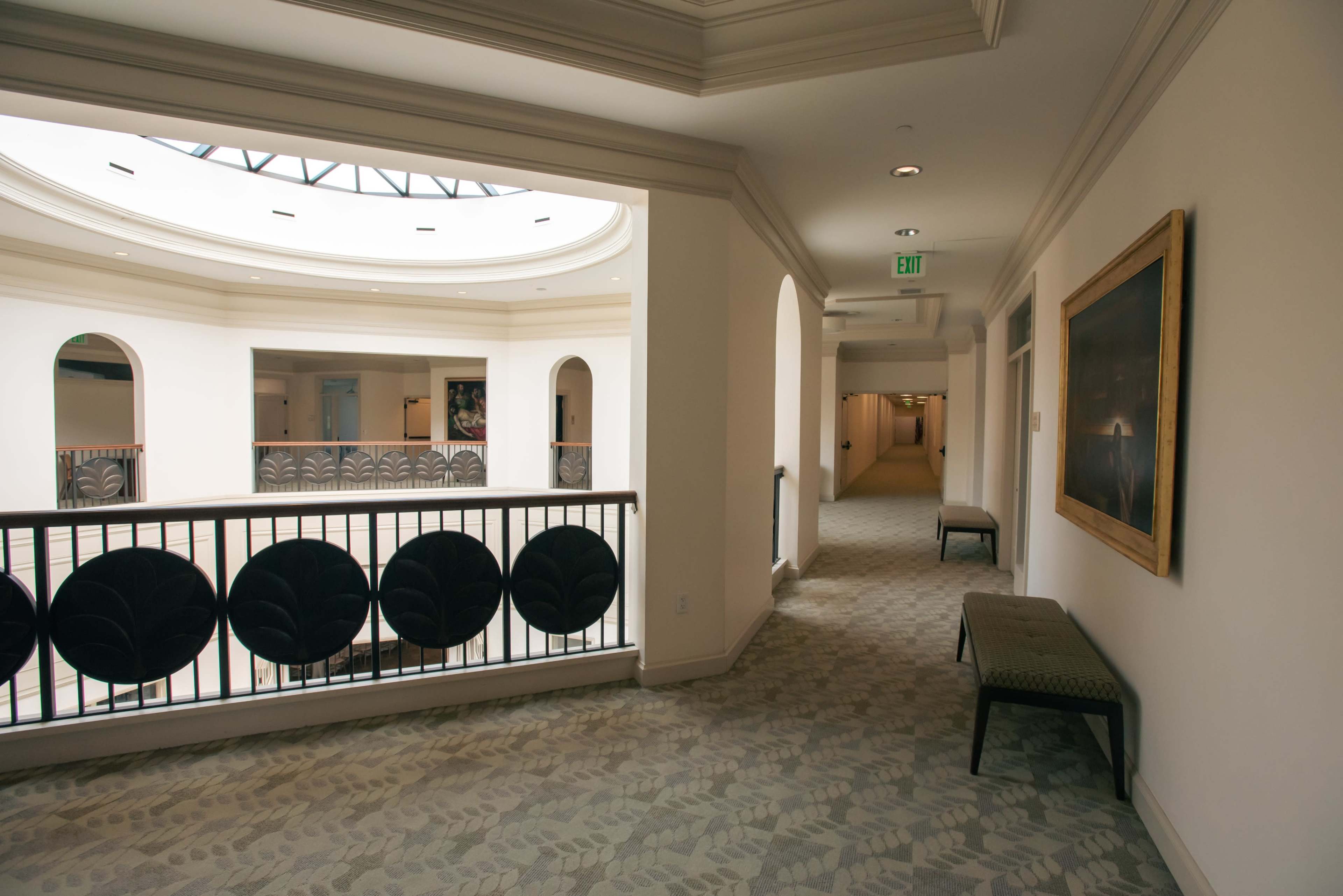 The image shows a well-lit corridor with a view of an atrium through arched openings, featuring benches along the walls and a railing with decorative elements.