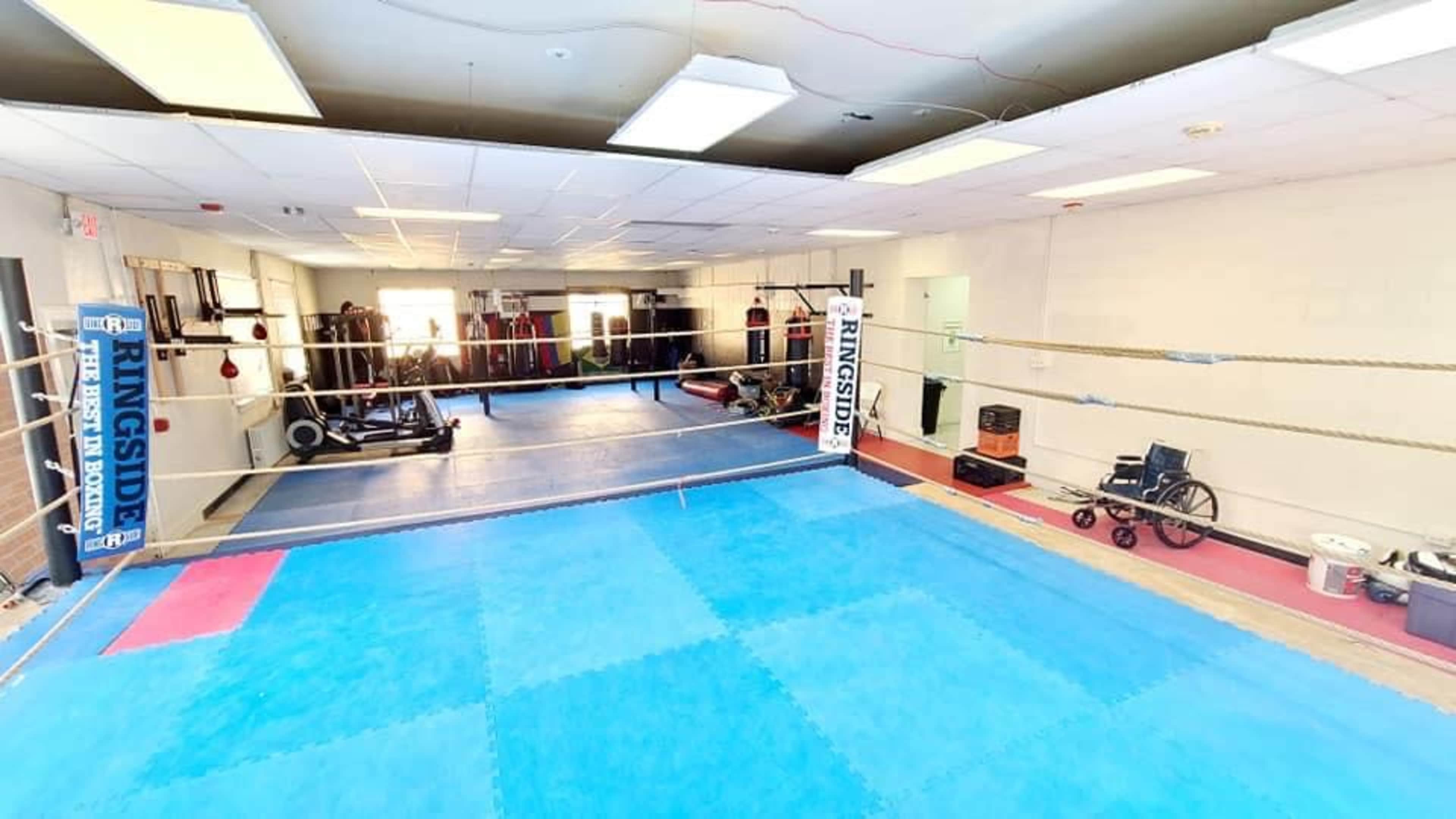 A well-lit boxing gym with a blue training mat and a boxing ring surrounded by various fitness equipment and training gear.