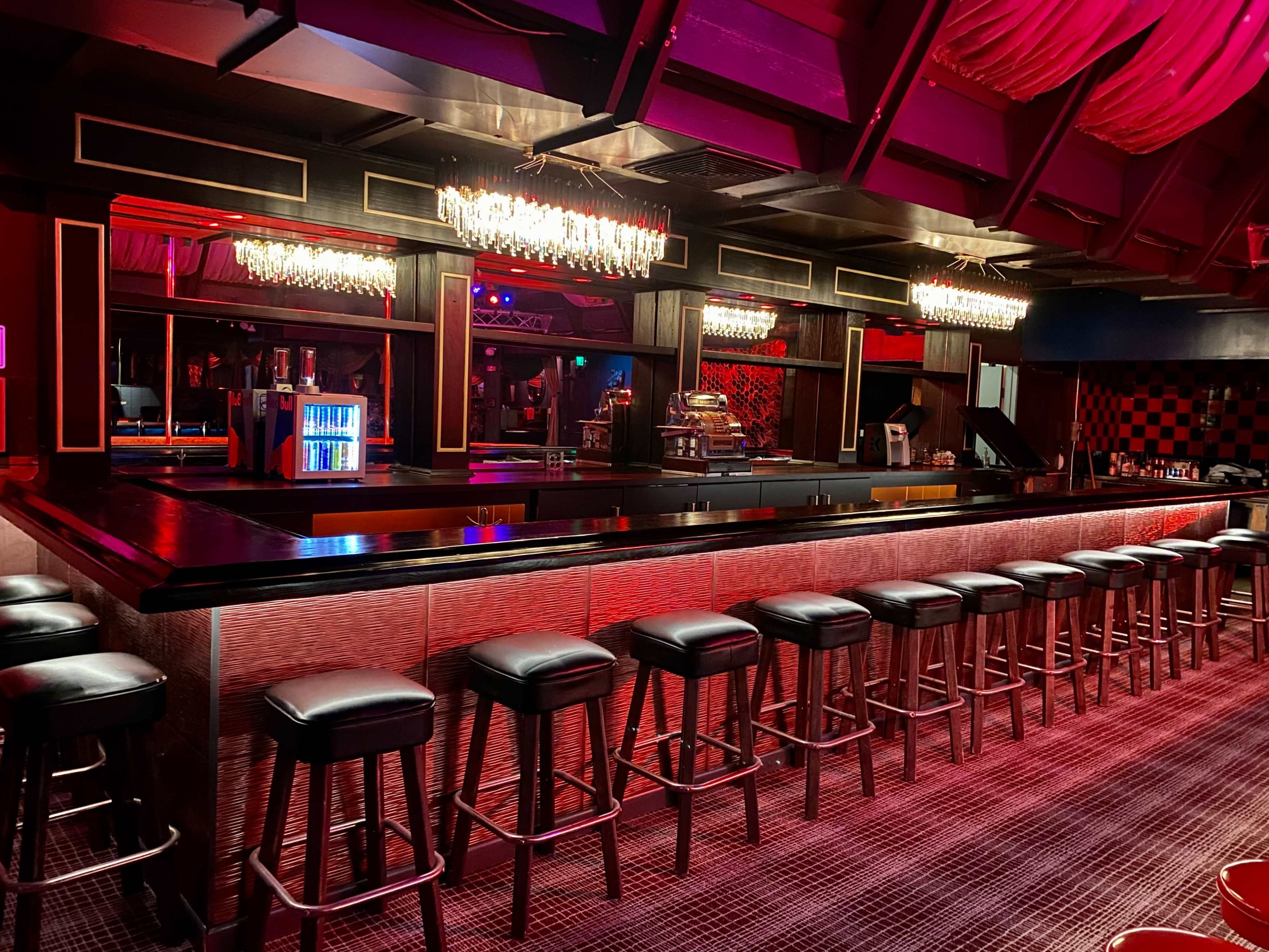 A dimly lit bar with a long counter, rows of bar stools, and crystal chandeliers overhead.