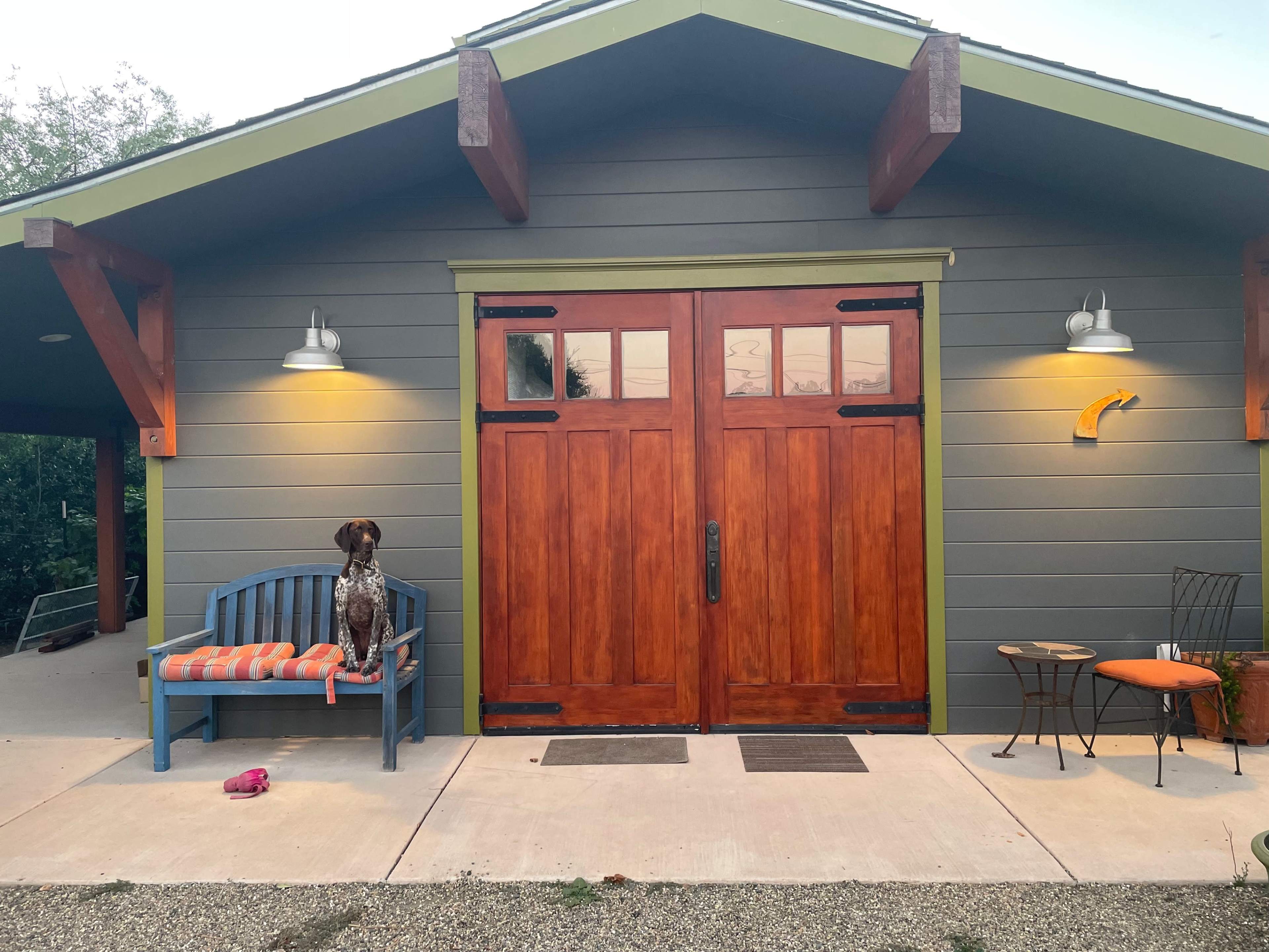 A dog sits on a blue bench in front of a large wooden door of a building with a gray exterior and two outdoor lights.