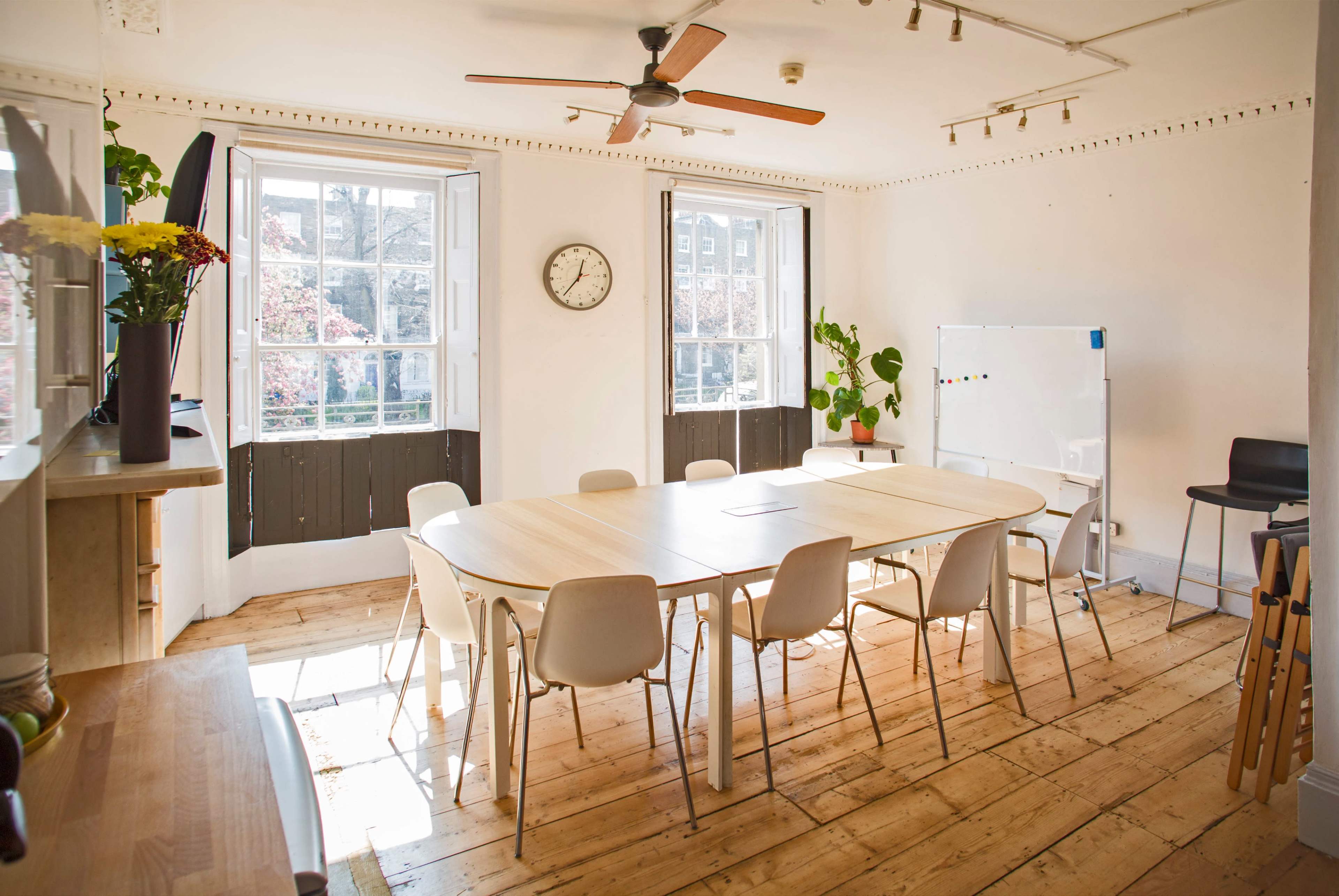 The image shows a bright meeting room with a large table surrounded by chairs, windows allowing natural light, and a clock on the wall.